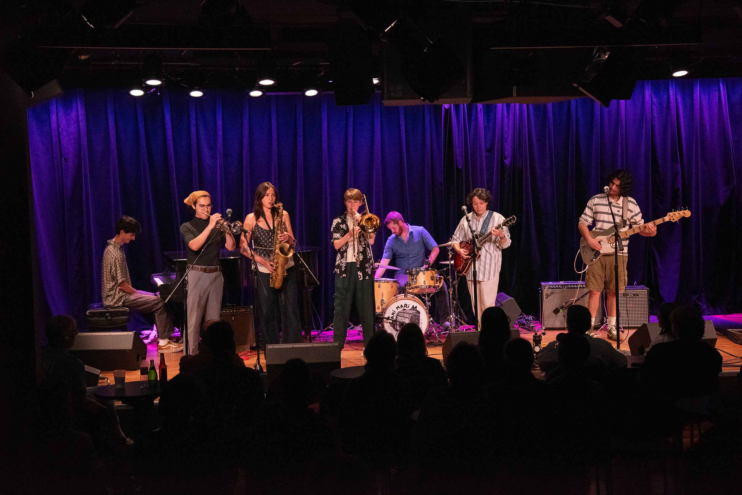 A seven-piece jazz band performs on a stage with blue-lit curtains in the background, and audience heads in the foreground.