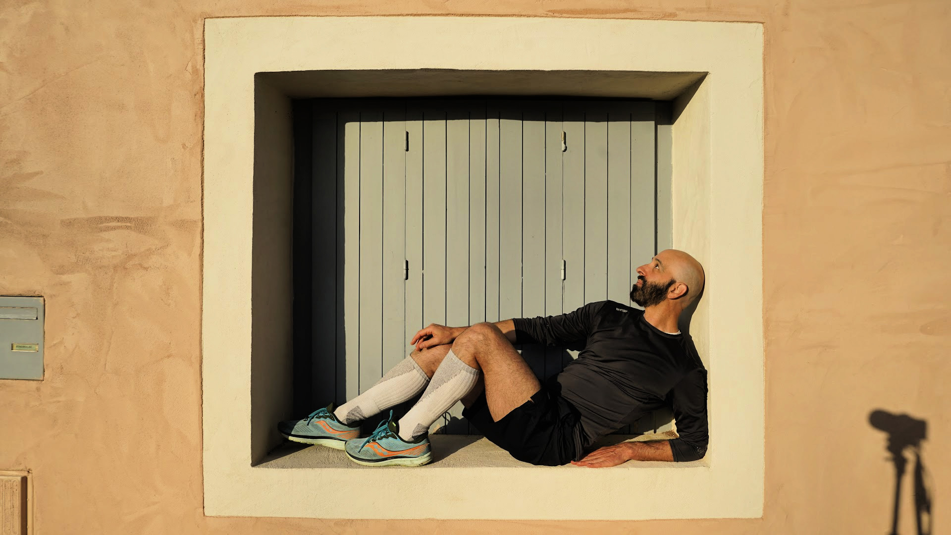 Daniel Gwirtzman poses leaning inside the window frame of a historic building with wooden shutters; attired in athletic clothing and sneakers.