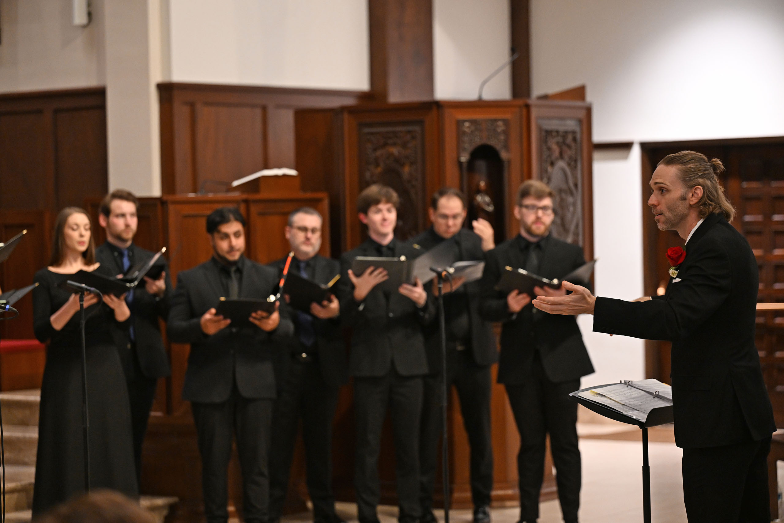 Daniel Knaggs conducts, with a line of seven singers visible in the background holding music.