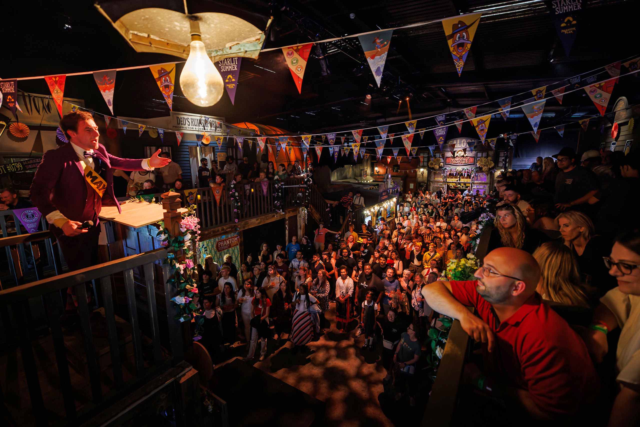 In a dark venue with signs and flags hanging all around, a costumed performer speaks from a balcony while a seated audience fills the space below and in the adjacent balcony.