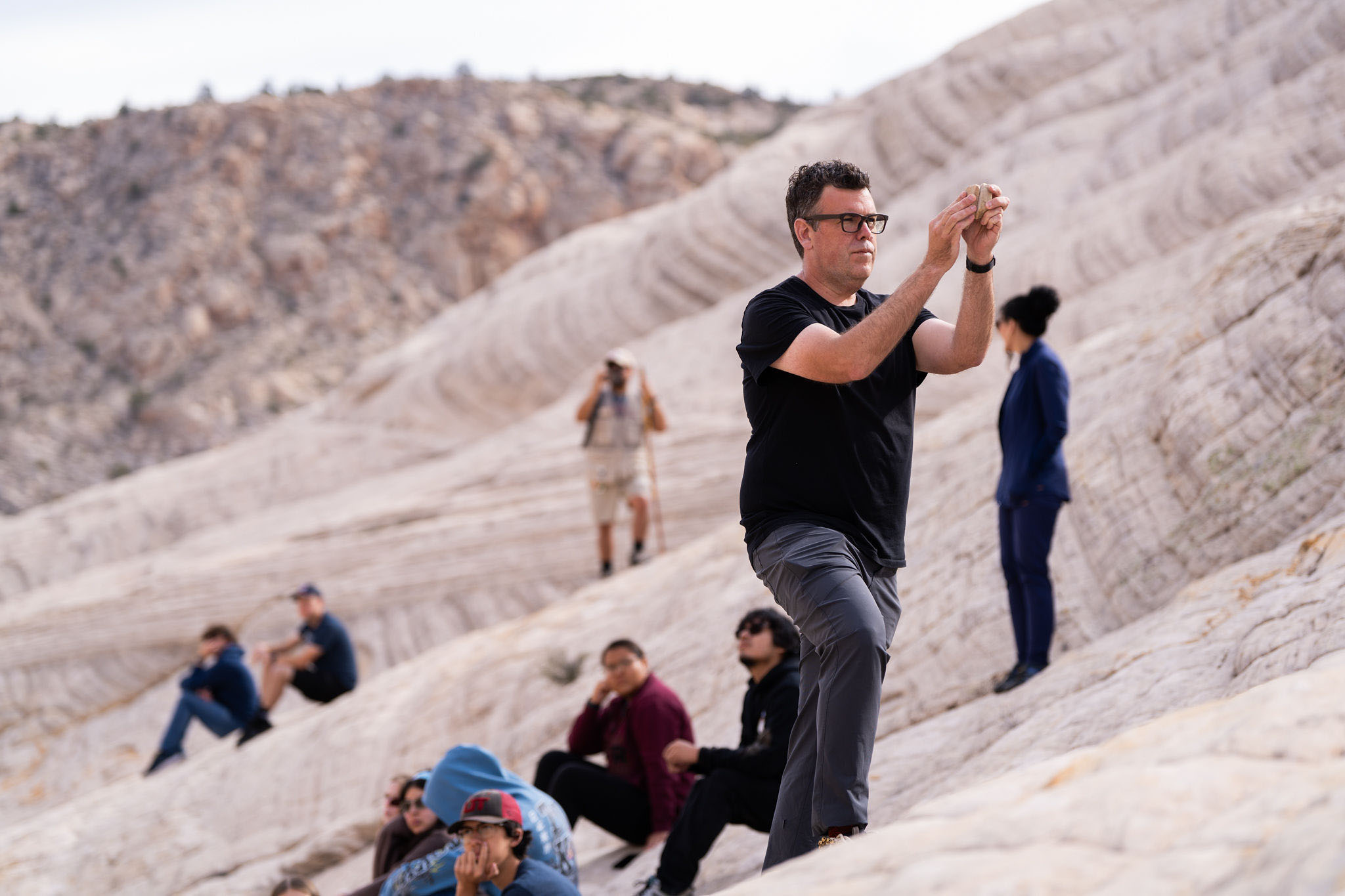 Candid photo of a group visiting a rocky rolling landscape, as Doug Perkins stands holding and examining a rock in his hands, with a group of others seated on the rock slope.