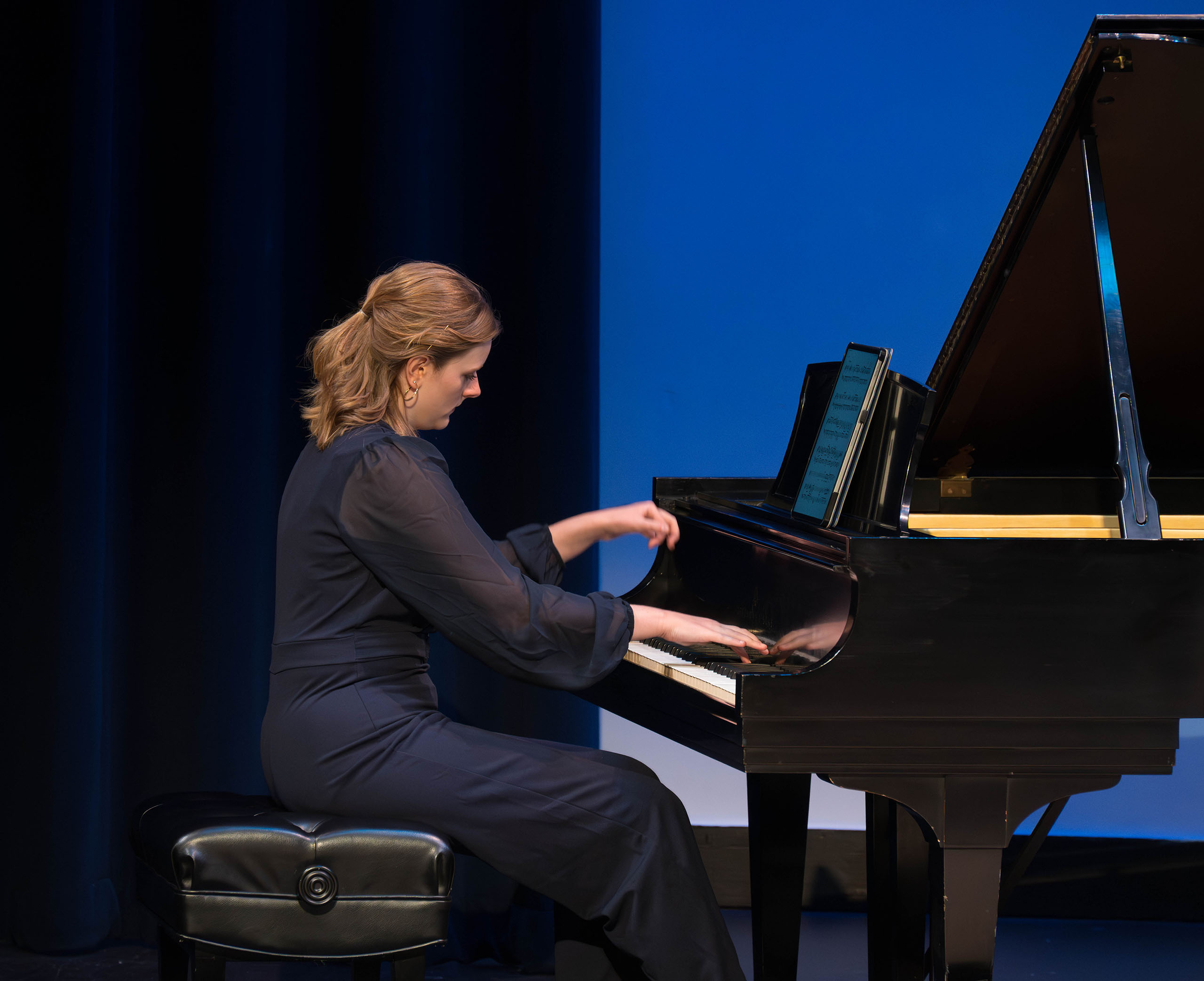 Eden West performs at a grand piano attired in a dark blue concert dress.