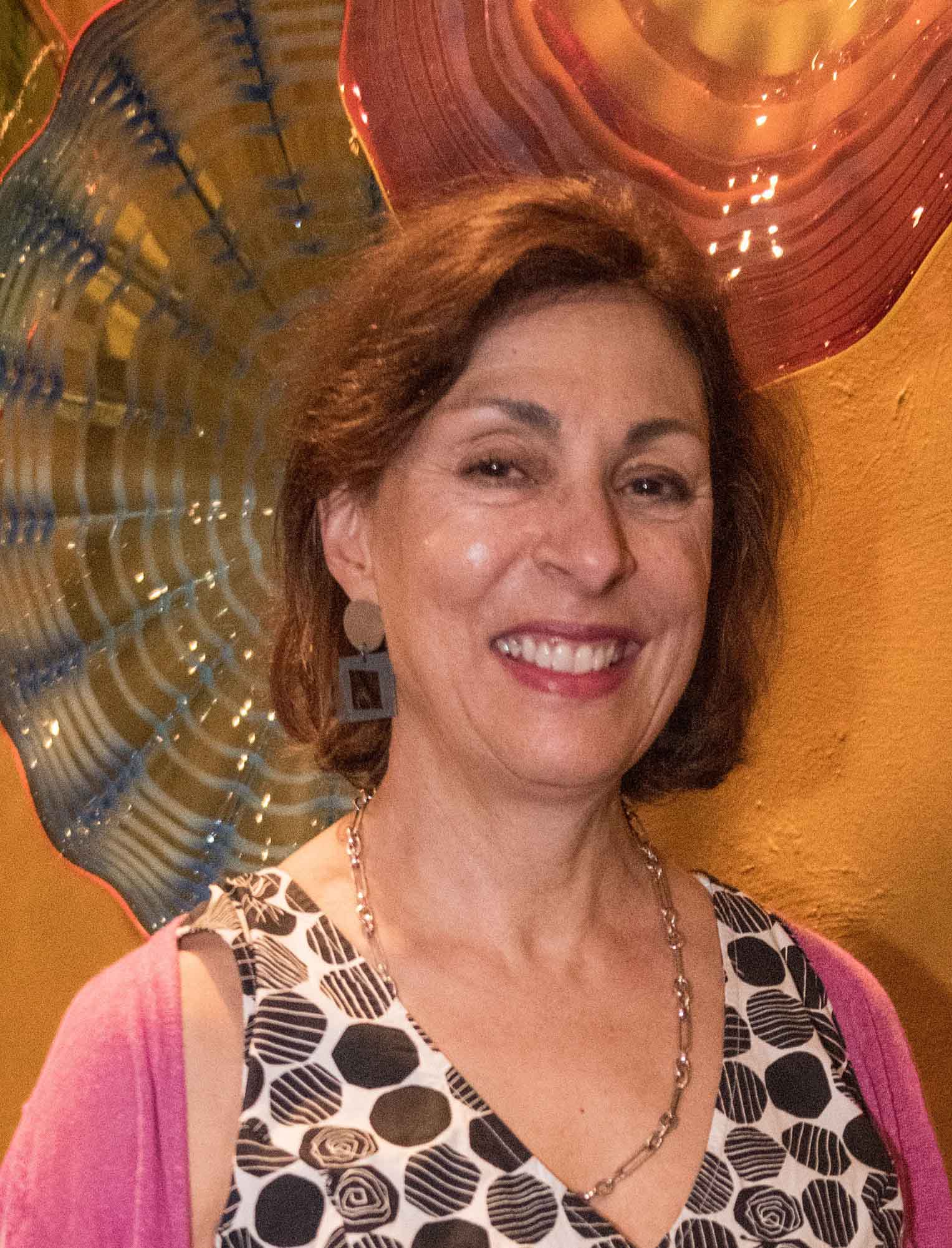 Elaine Guregian poses standing in front of two blown glass sculptures on a wall.
