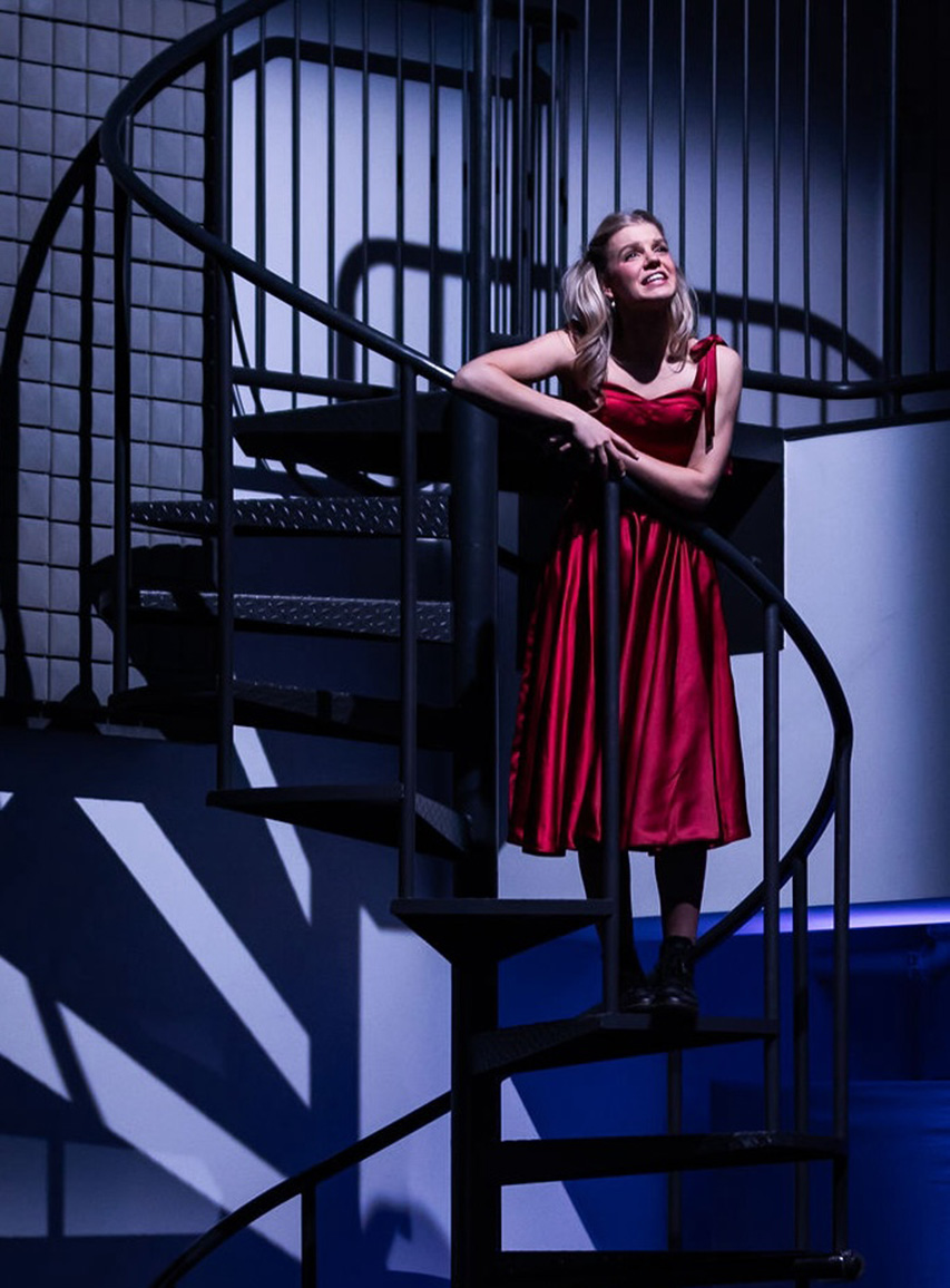 Ella Olesen performs on stage, standing on a spiral staircase and costumed in a red dress.