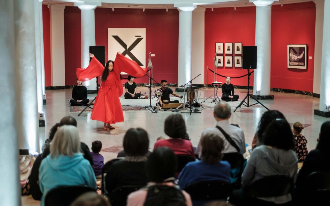 Fangfei Miao performs dance in a flowing red costume in the Apse at the U-M Museum of Art accompanied by musicians, with an audience seated in the foreground.