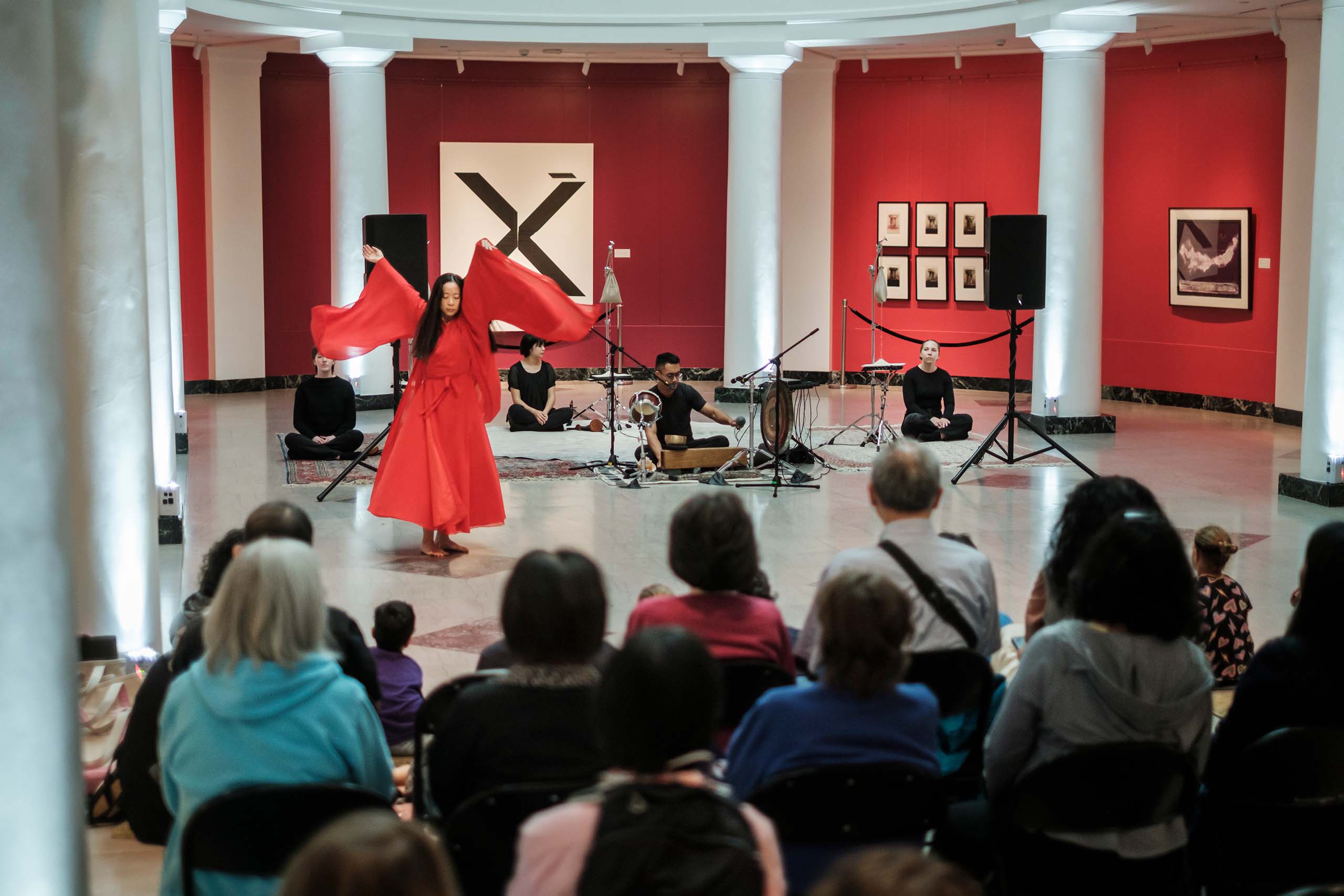 Fangfei Miao performs dance in a flowing red costume in the Apse at the U-M Museum of Art accompanied by musicians, with an audience seated in the foreground.