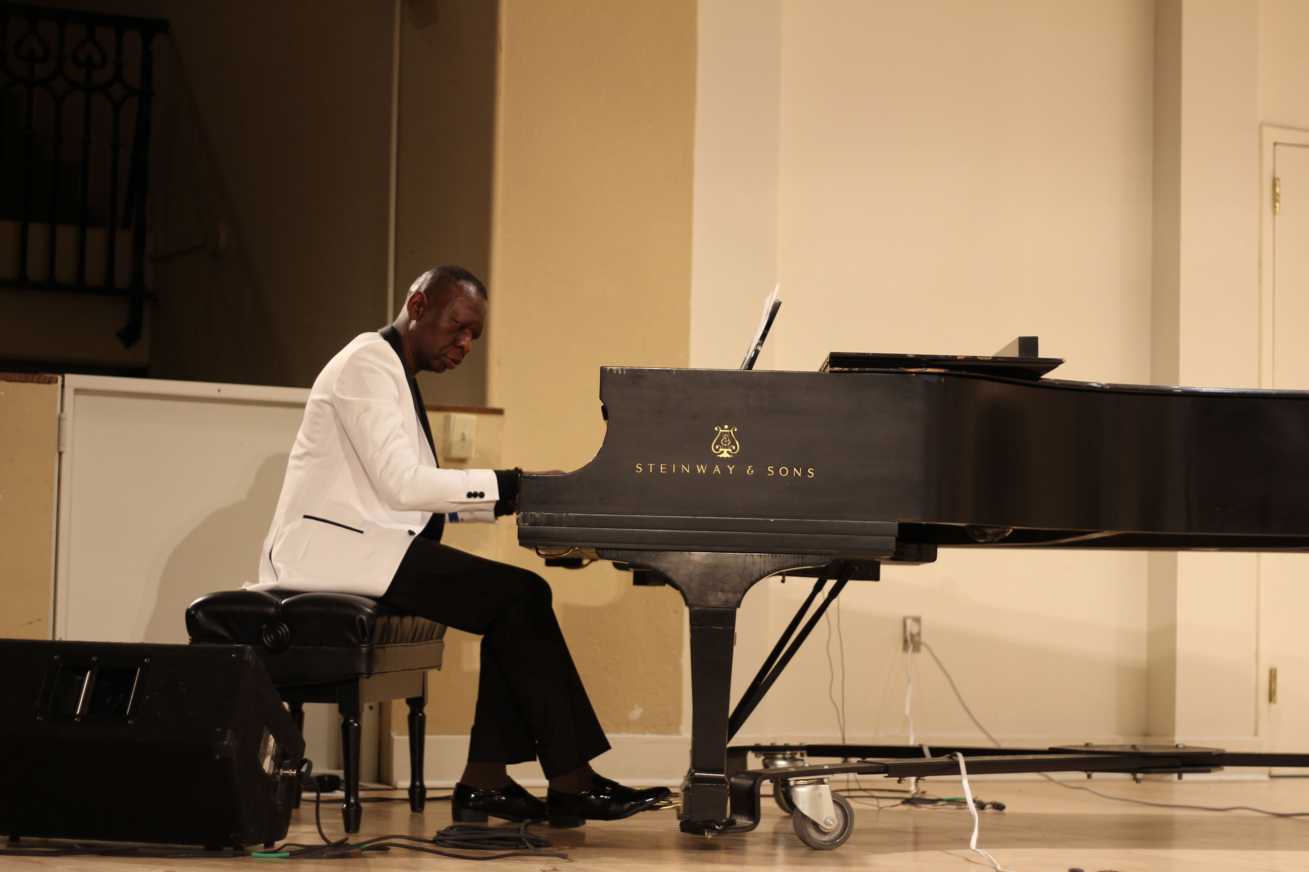 Godfrey Lubuulwa performs on a grand piano, attired in a white tuxedo coat.