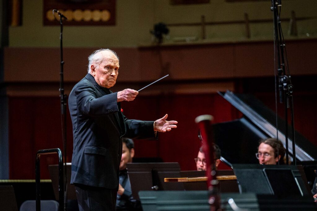 H. Robert Reynolds conducts a performance with arms raised, holding a baton, attired in formal black. A piano, wind musicians, and music stands are visible from the ensemble surrounding him.