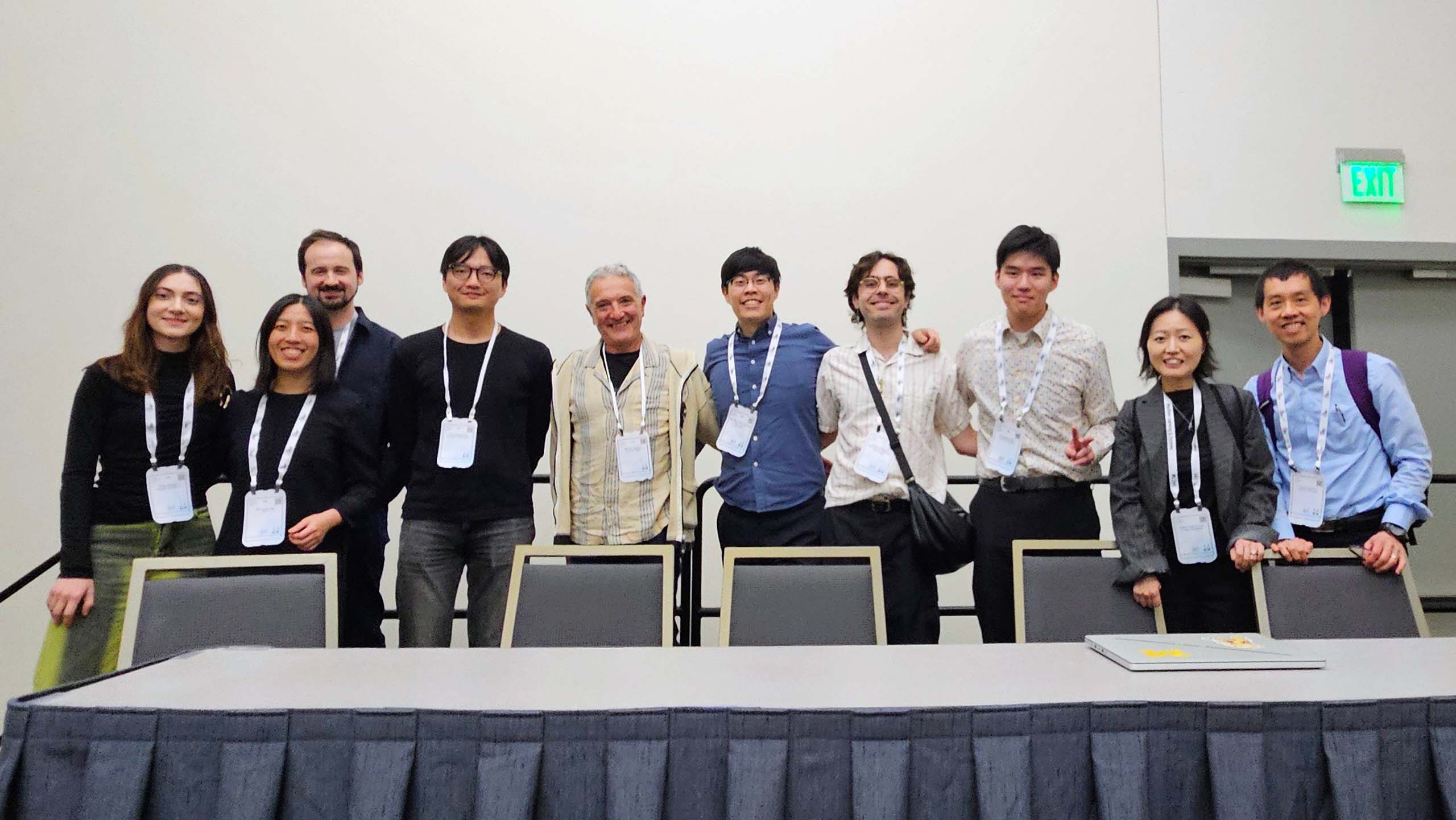 Ten people pose standing behind a panelist's table, all wearing hanging conference nametags.