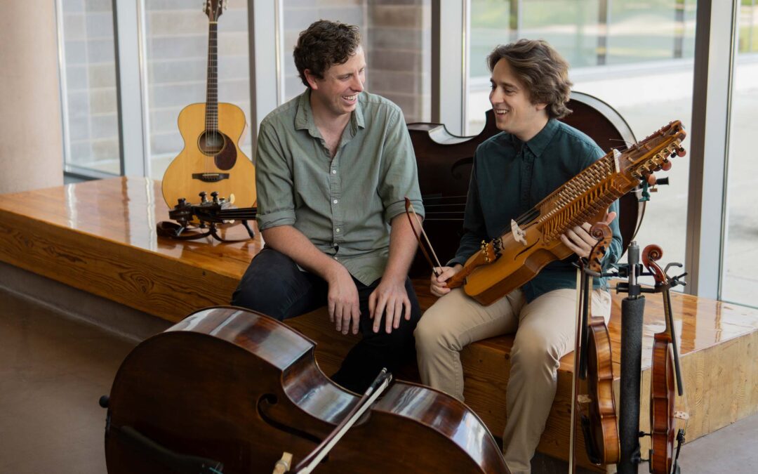 Candid photo of Jacob Warren and Grant Flick seated together in a lobby space, surrounded by six string instruments of various sizes.