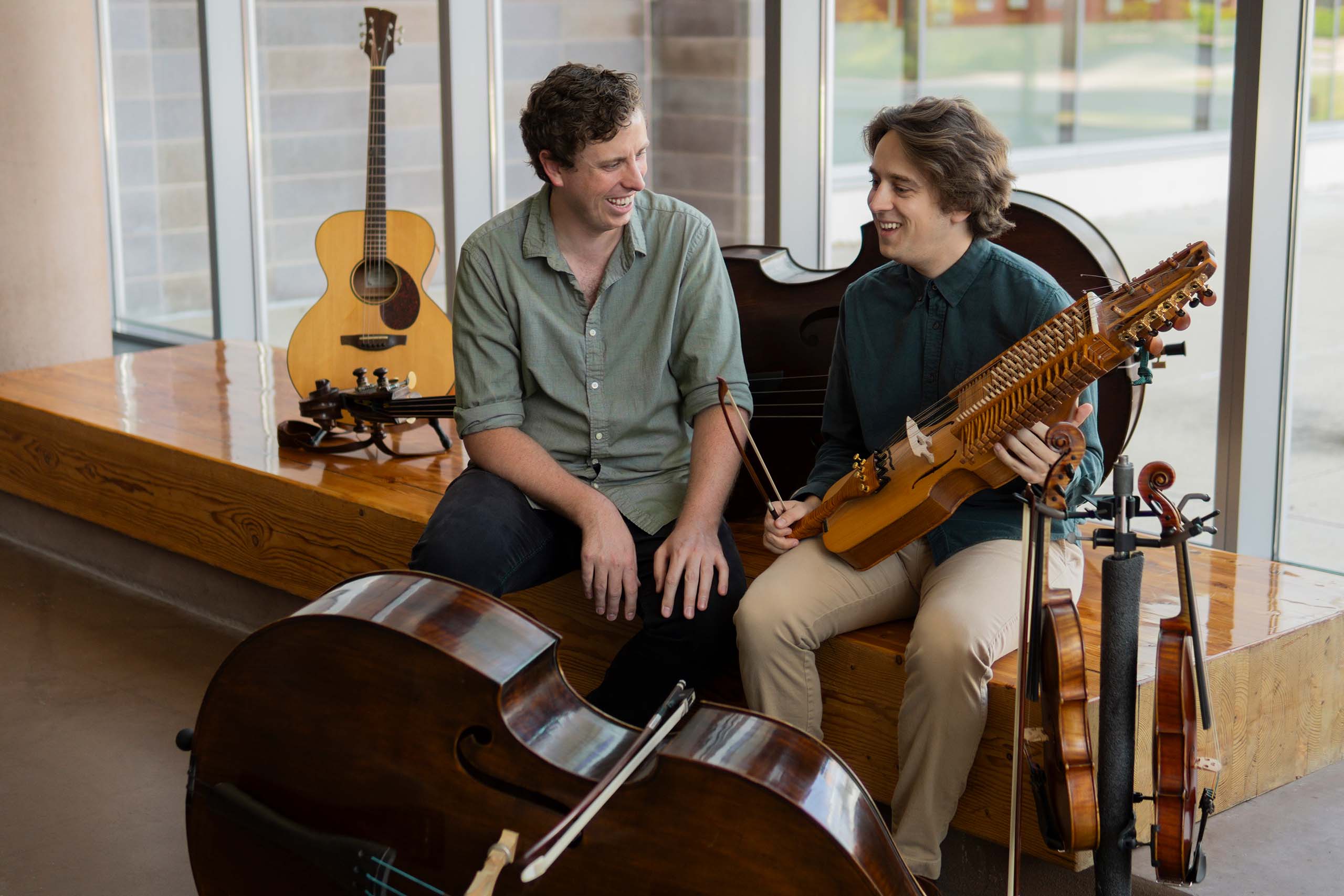 Candid photo of Jacob Warren and Grant Flick seated together in a lobby space, surrounded by six string instruments of various sizes.