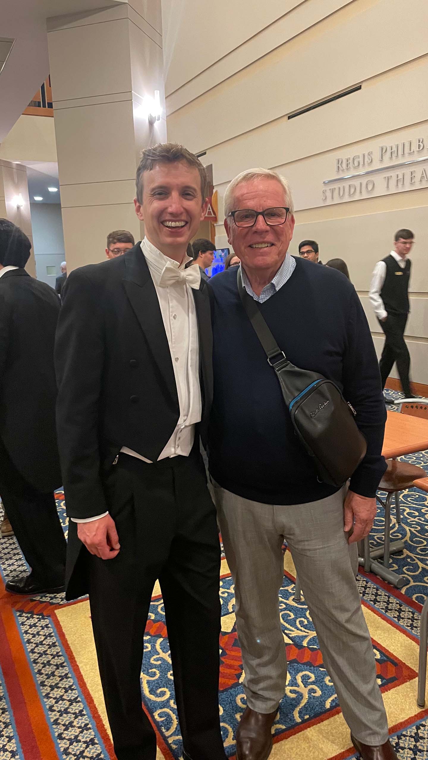 Two people pose standing in a venue lobby, with one in formal concert attire.