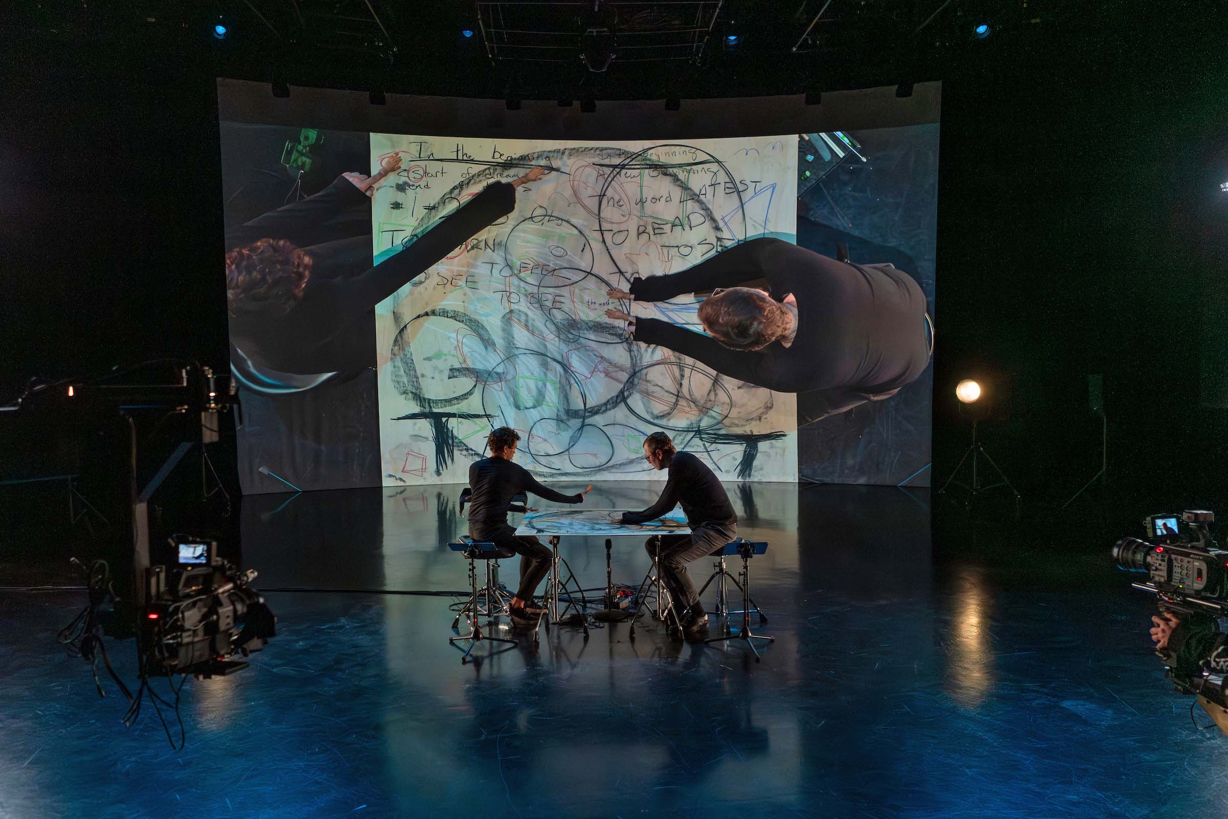 In a dark recording studio, two people sit at a table attired in black. A large projection behind them displays the letters and scribbles shapes on the table they sit at.