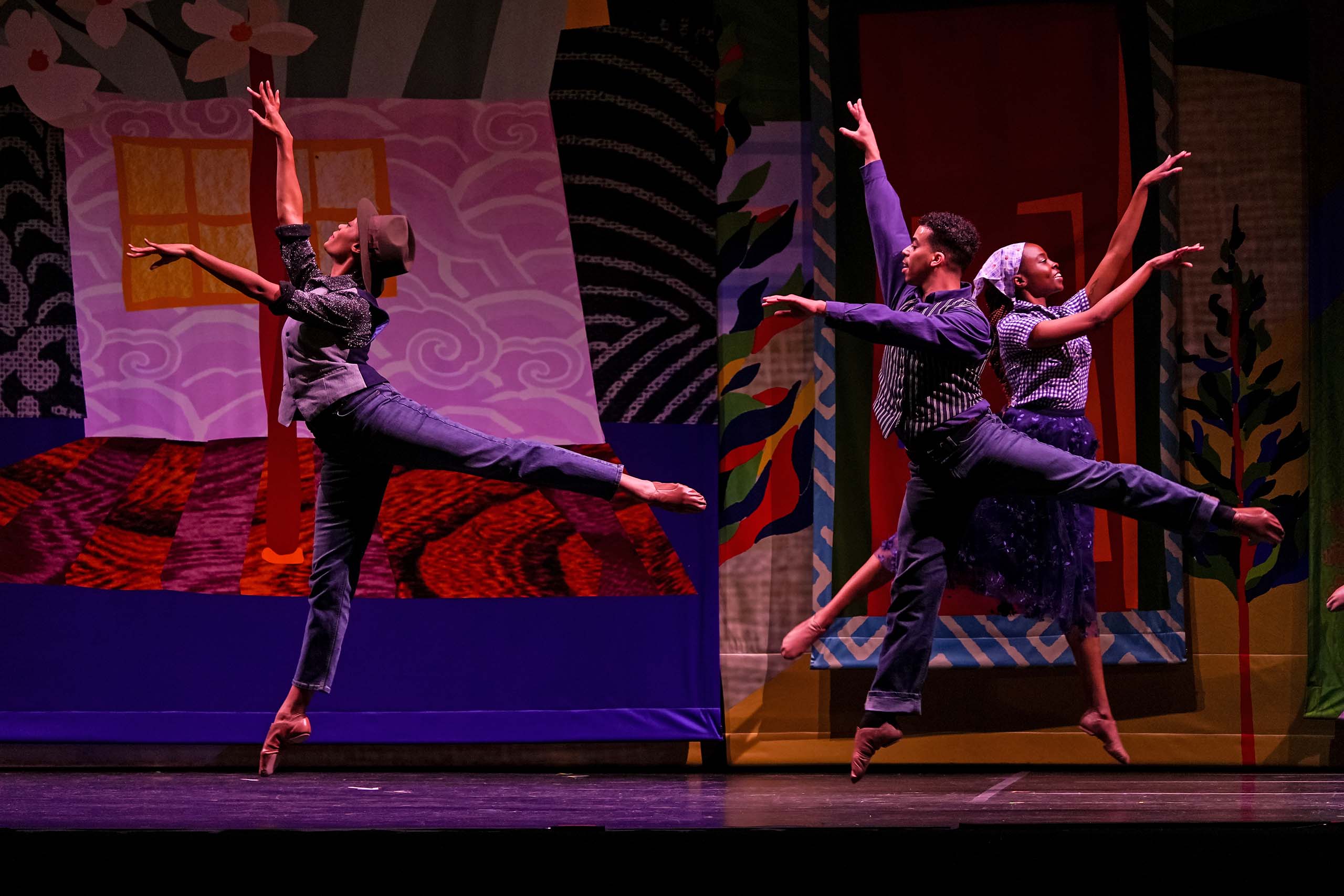Three dancers perform leaps on stage, costumed in dark blue pants and vests, with patterned cloths of the costumes and set evoking a mid-20th century domestic scene.