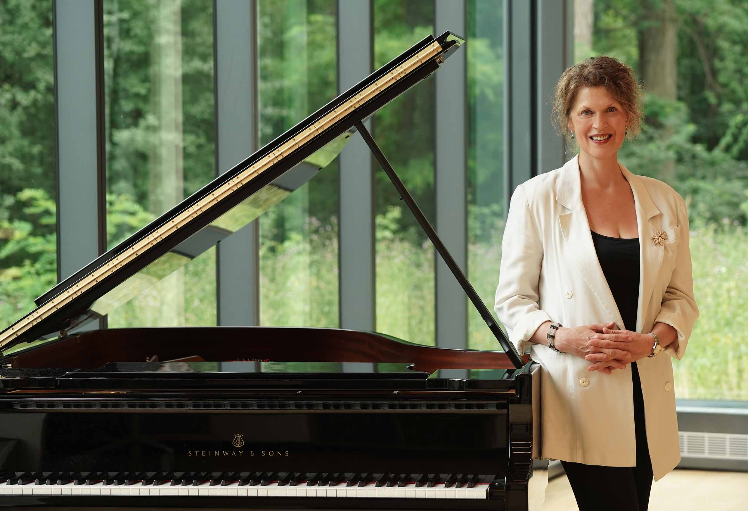 Kathryn Goodson poses standing next to a grand piano in a dance studio with large window walls overlooking green space.