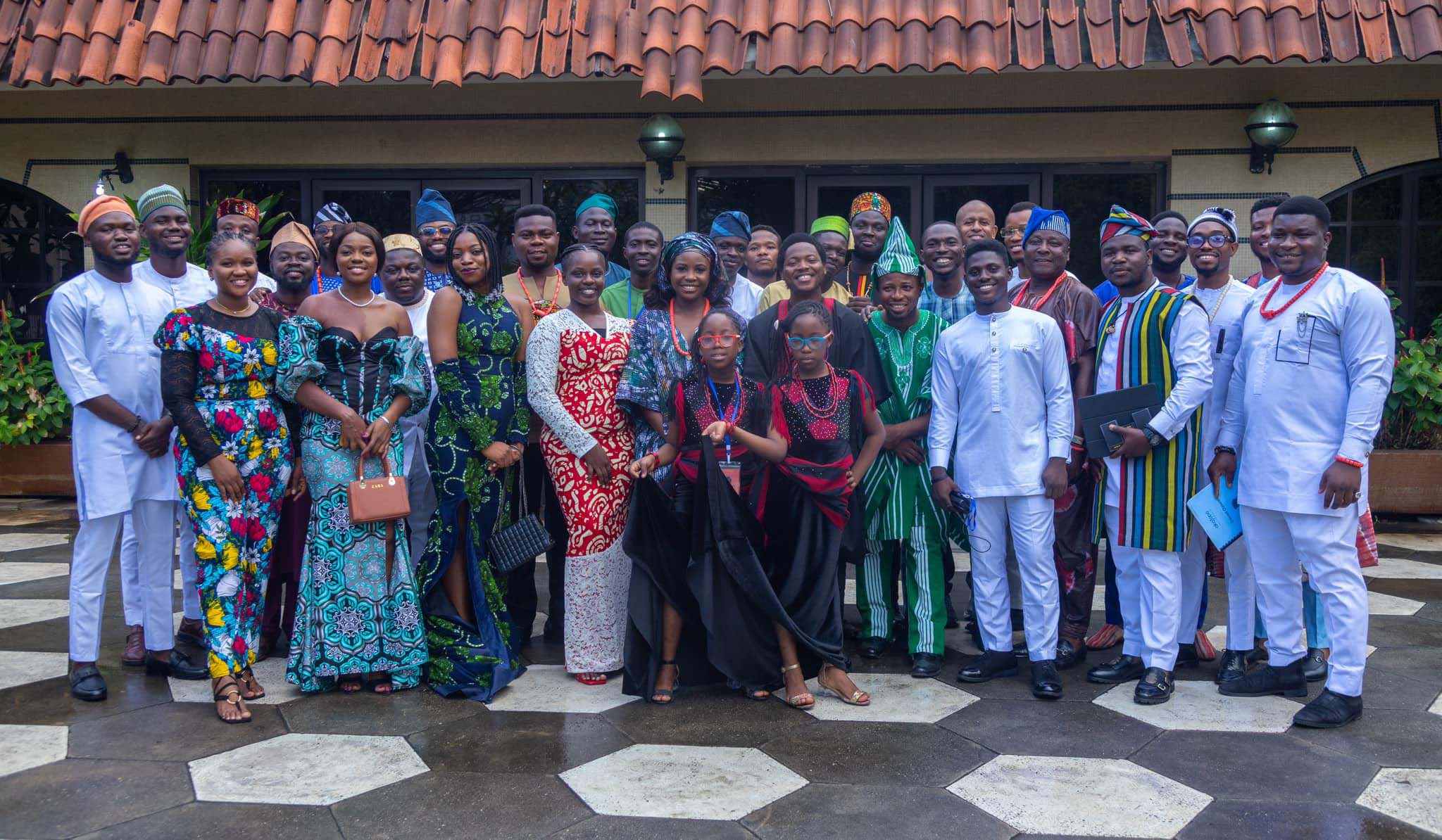 About 3 dozen people stand posing together in a courtyard, attired in formal African clothing.