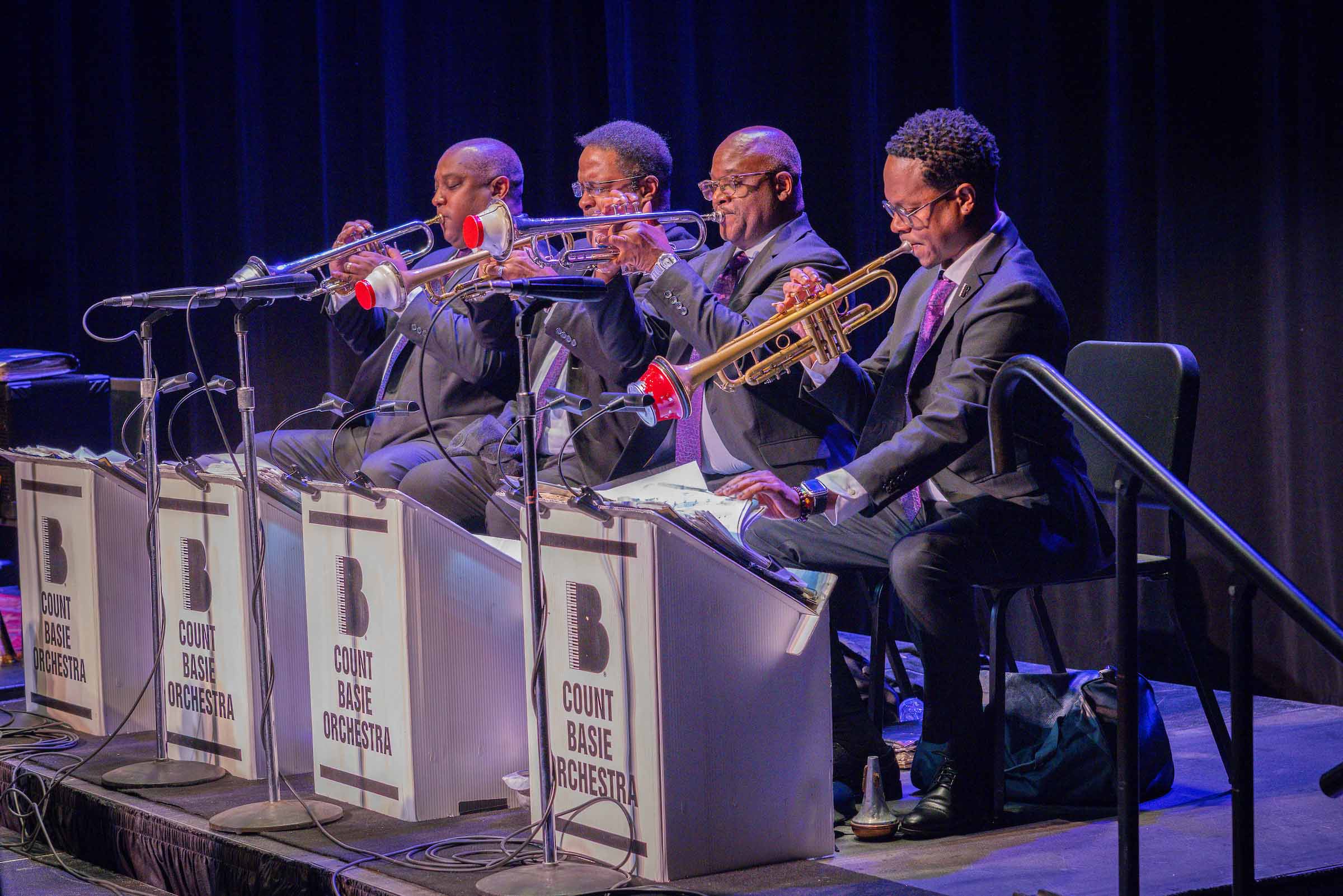Four jazz trumpters perform seated on stage with trumpet mutes, facing their music on matching lecterns displaying the Count Basie Orchestra logo.