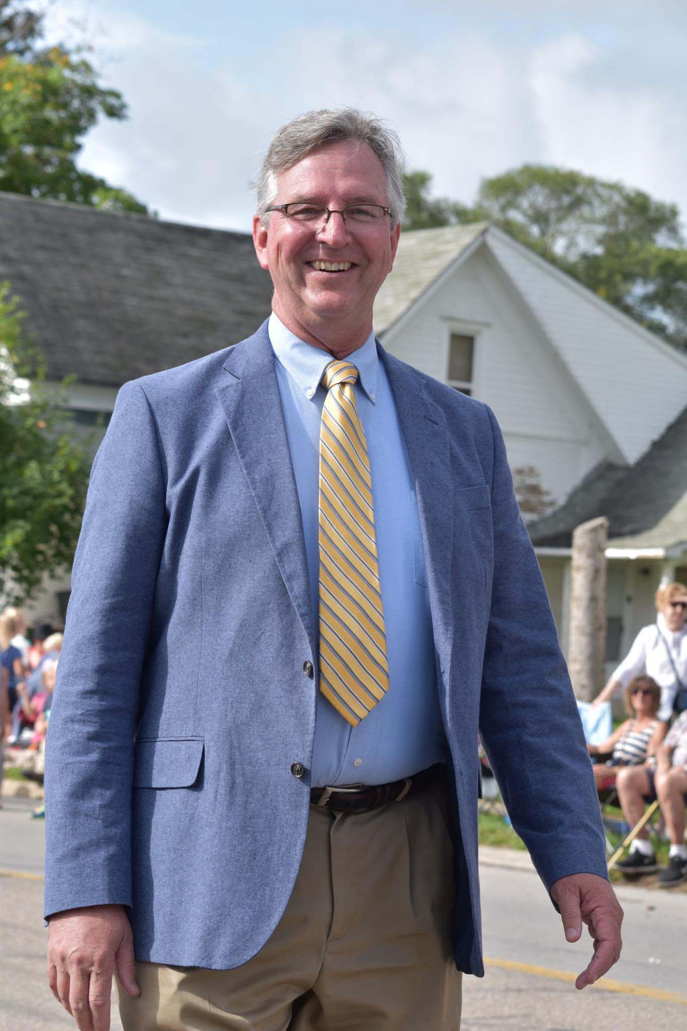Mike Filkins stands outdoors on a town street, attired in a suit, with parade viewers seated along the street in the background.