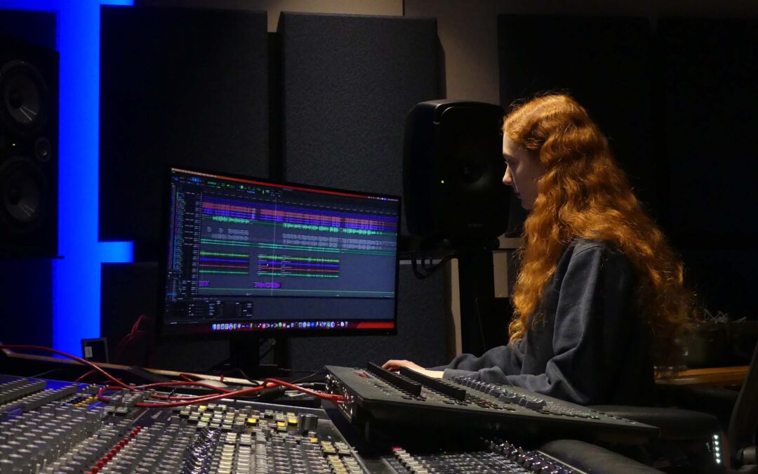 Renata Schmult works in music software on a computer screen near a mixing board.