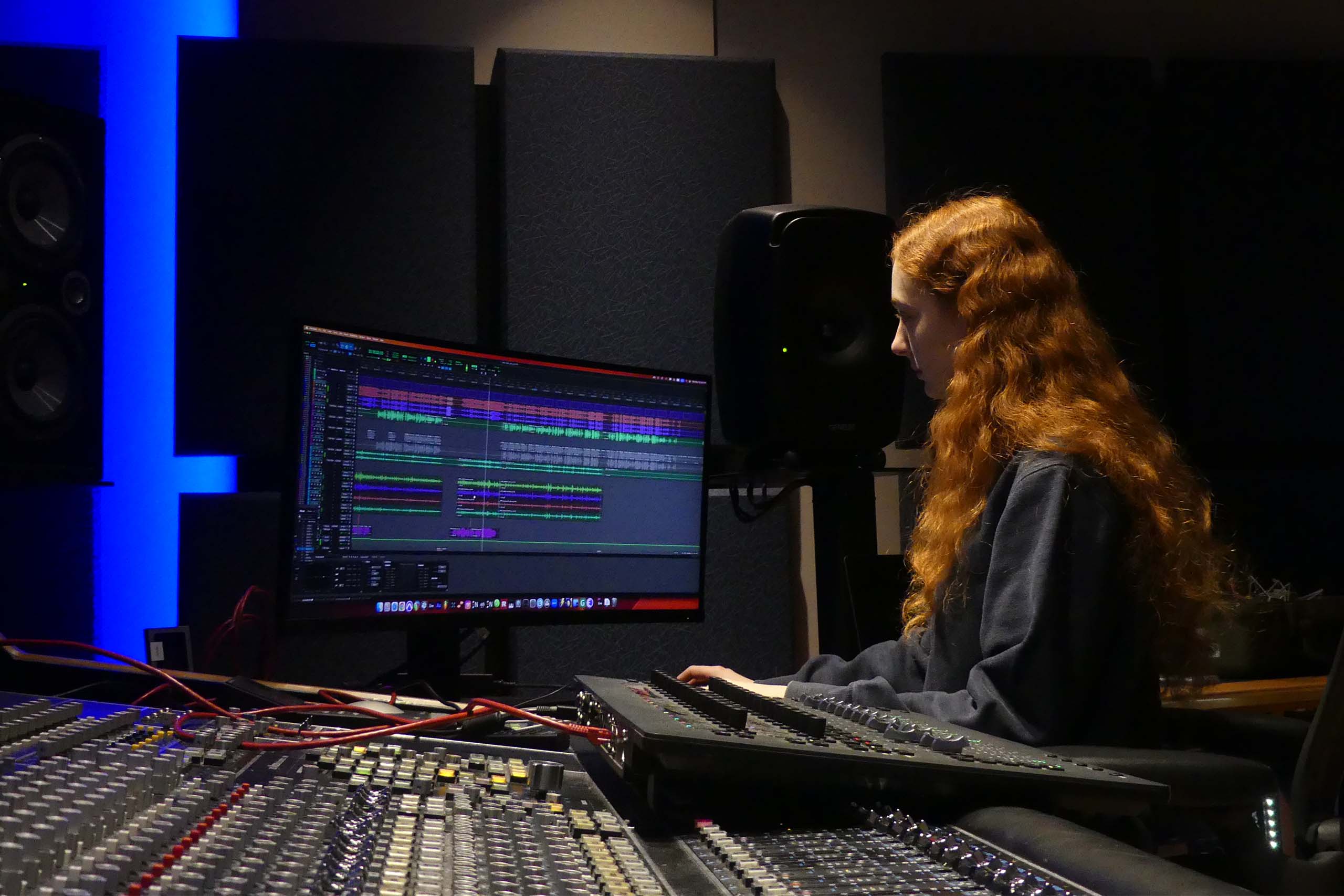 Renata Schmult works in music software on a computer screen near a mixing board.