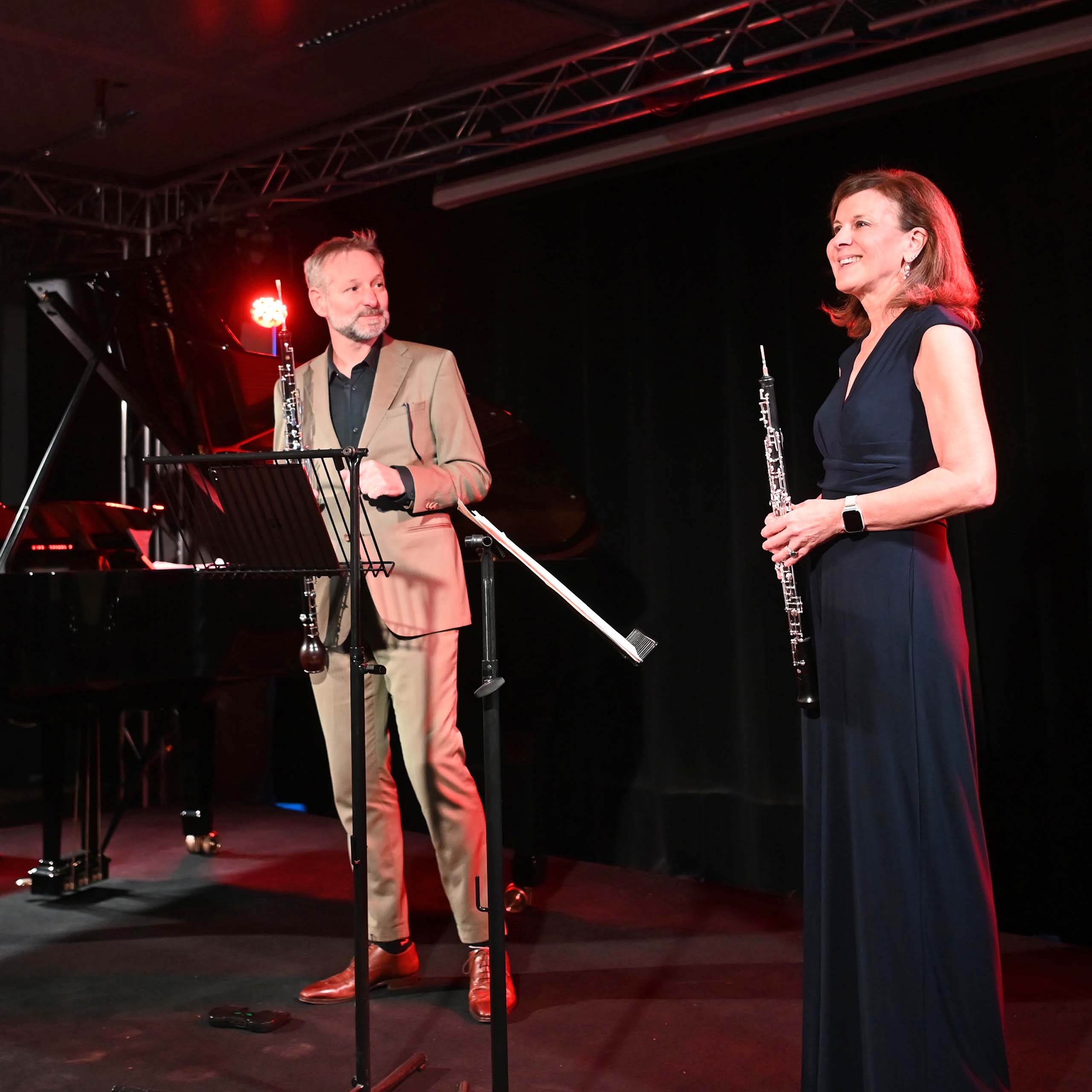 Two performers holding oboes stand on a dark stage in concert attire.