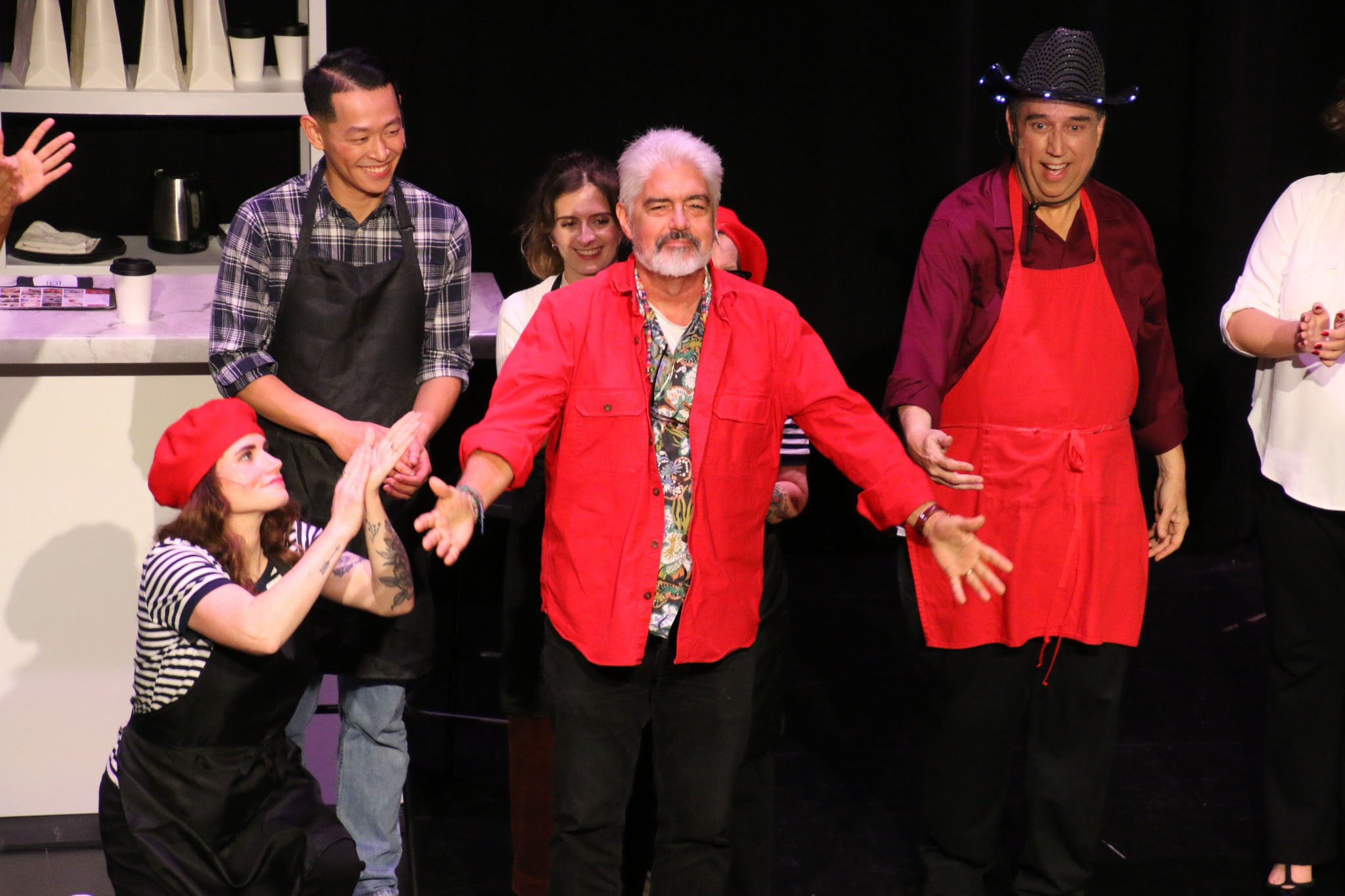 Neal Learner bows on stage, as performers costumed in aprons clap alongside him.