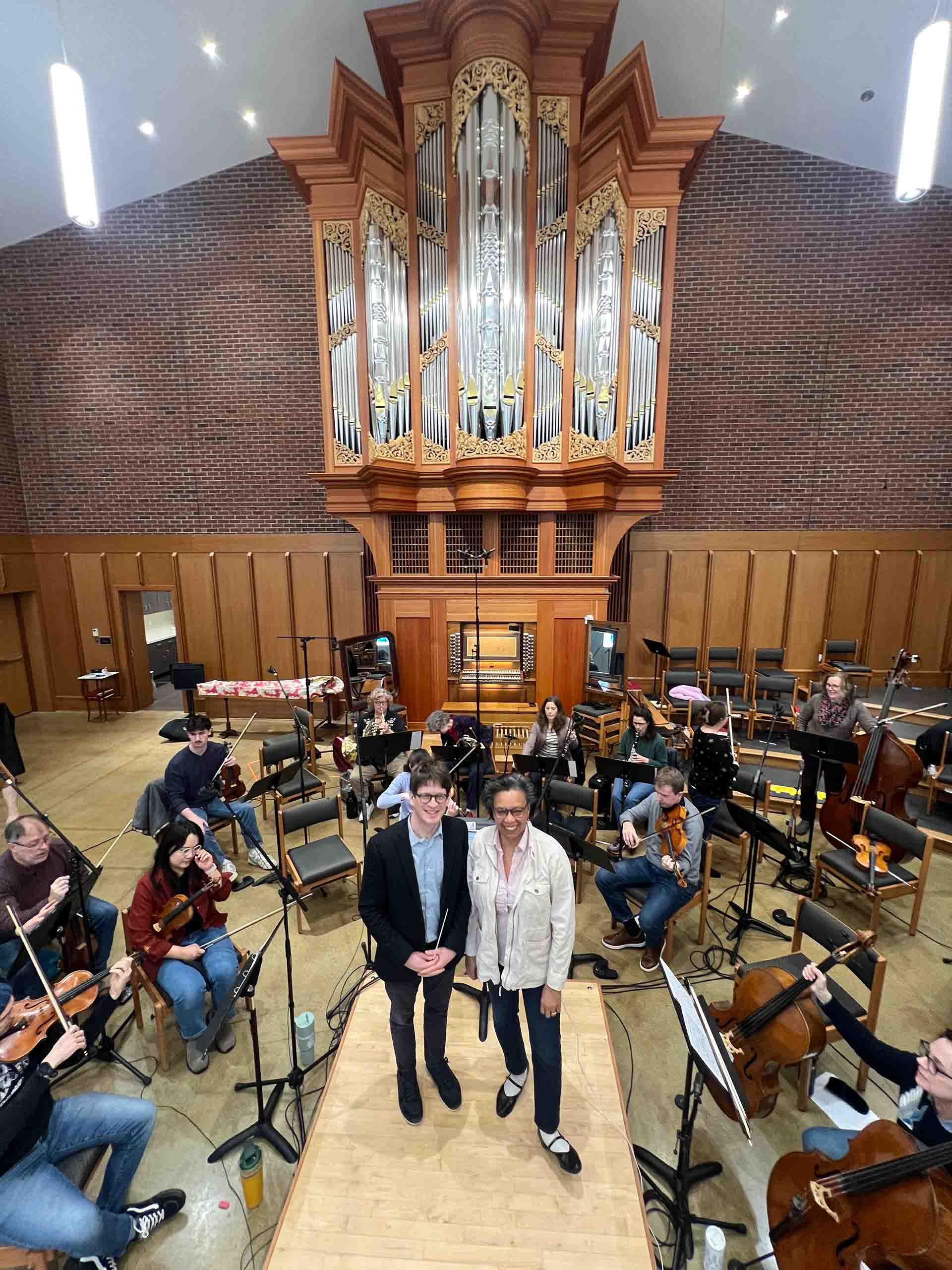 Two people pose standing in a venue with high ceilings and an organ, surrounded by a small orchestra doing rehearsal warm-ups.