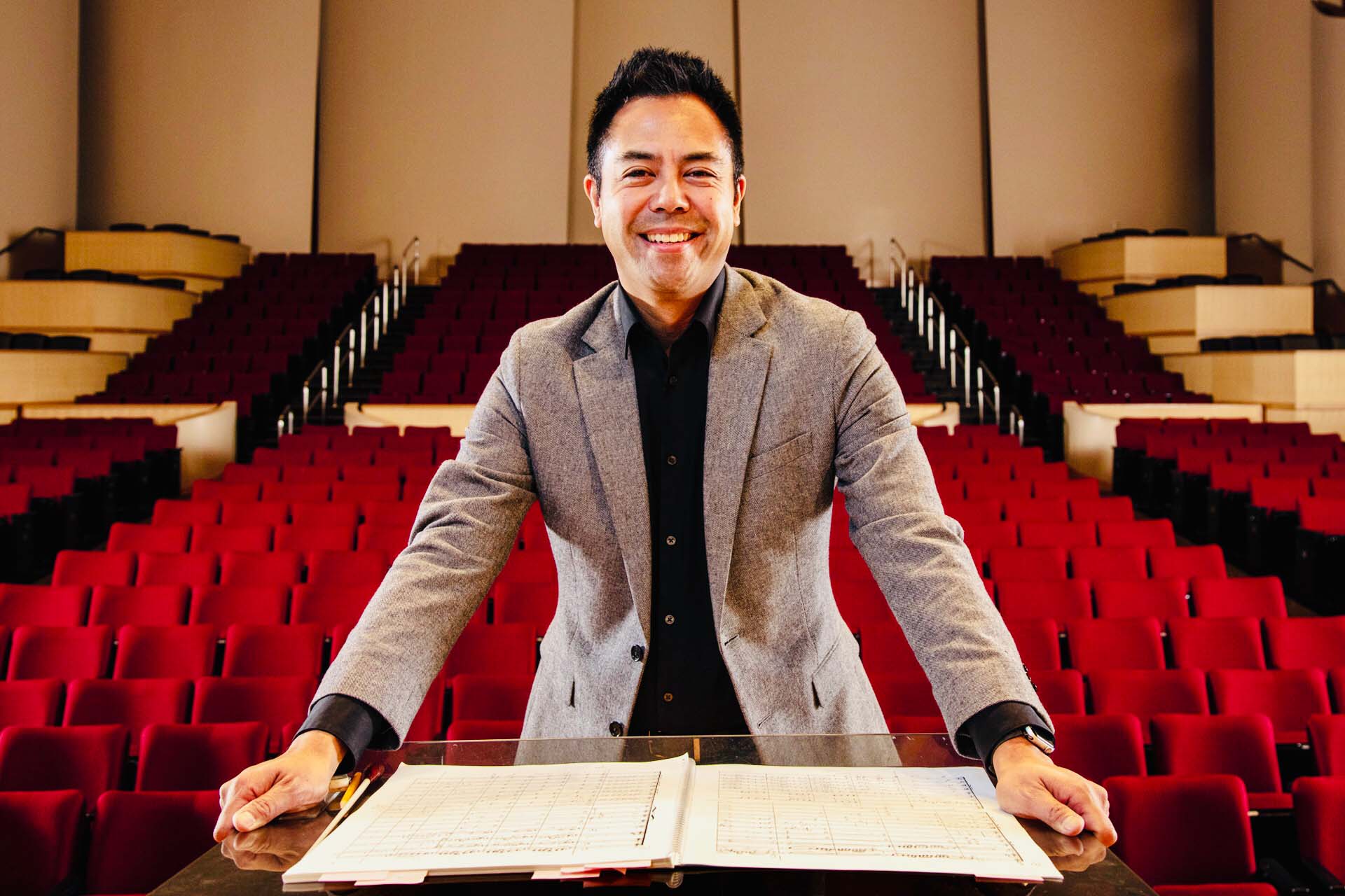 Nikolas Caoile poses standing at an open musical score, with an empty theatre house with red seats in the background.