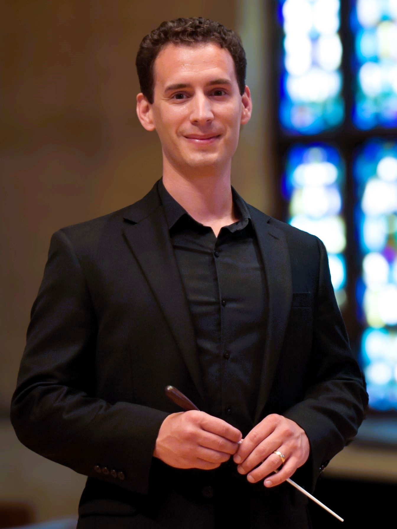 Portrait of Peter Kadeli in concert attire, holding a conducting baton, with stain glass windows in the background.
