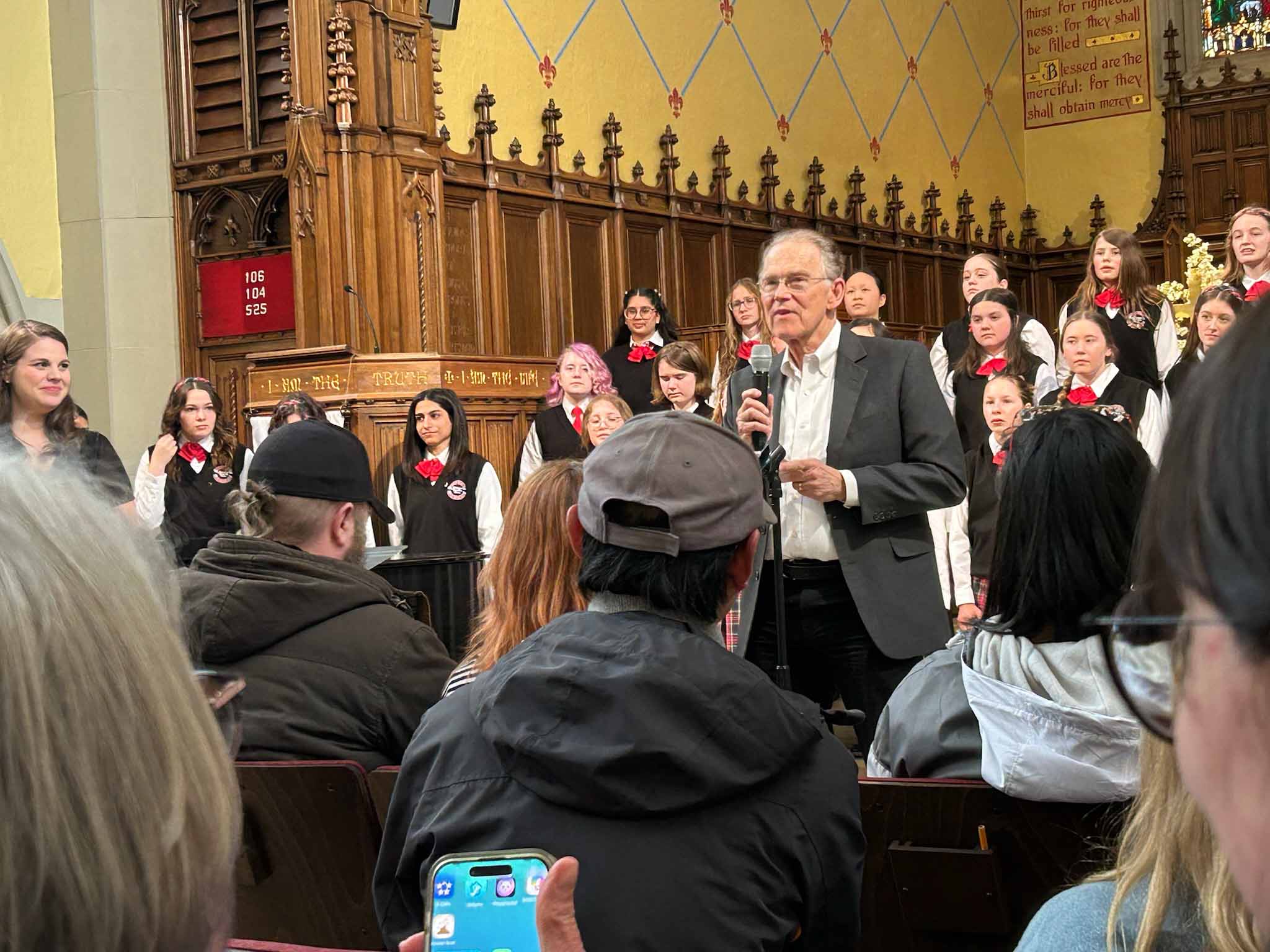 In a church venue, Richard Jennings speaks into a microphone, with a children's choir in the background and seated audience in the foreground.
