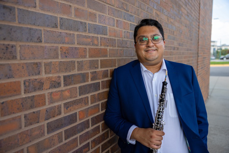 Portrait of Sagar Anupindi standing outdoors by a brick wall, holding his oboe.