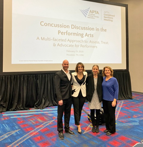 Four people pose standing in a convention space, in front of a slide projection with the title "Concussion Discussion in the Performing Arts"