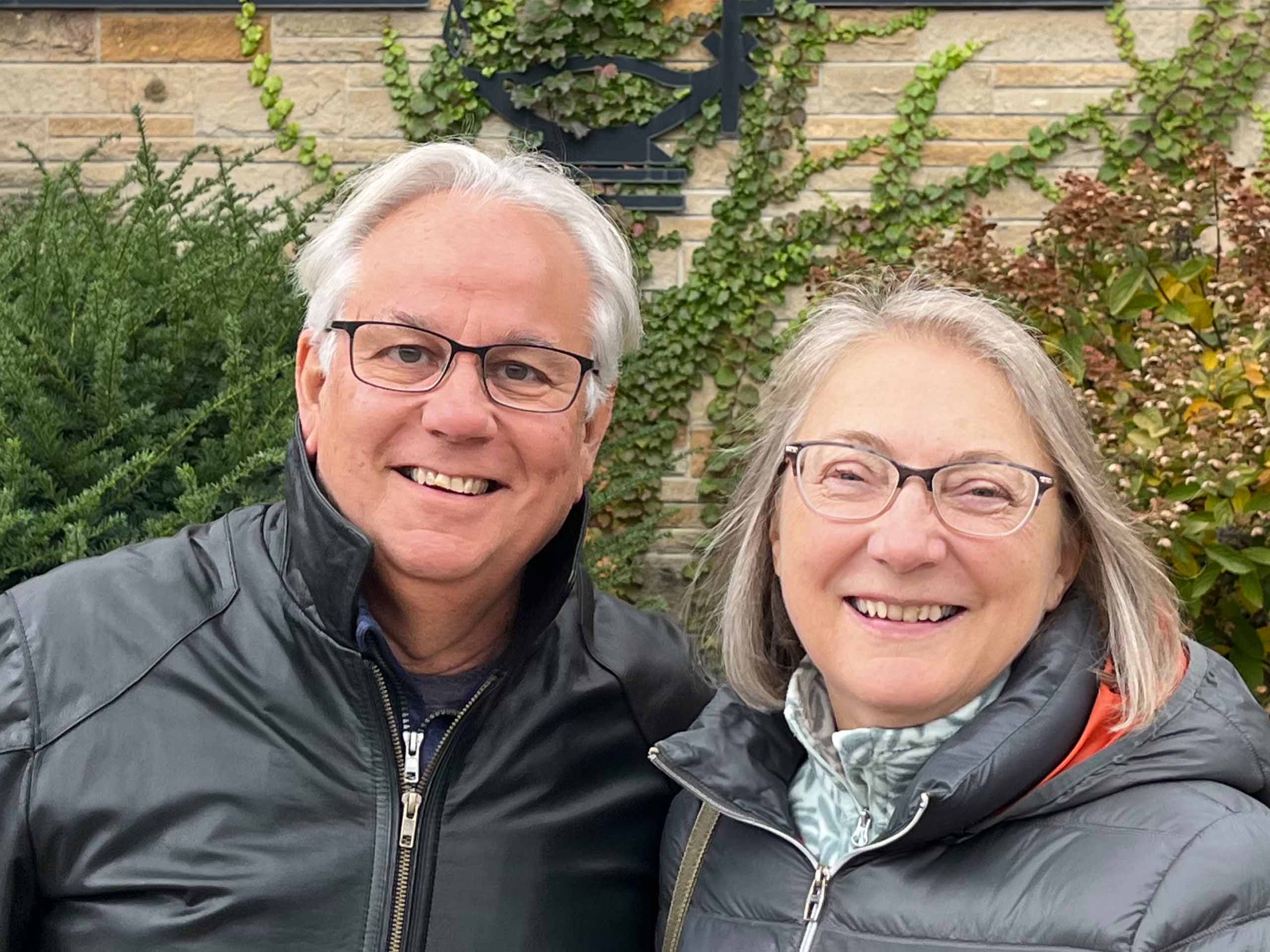 Sonya Szabo-Reynolds with her husband Patrick Reynolds Sonya Szabo-Reynolds and Patrick Reynolds pose standing together wearing coats outdoors, with a brick wall and foliage in the background.