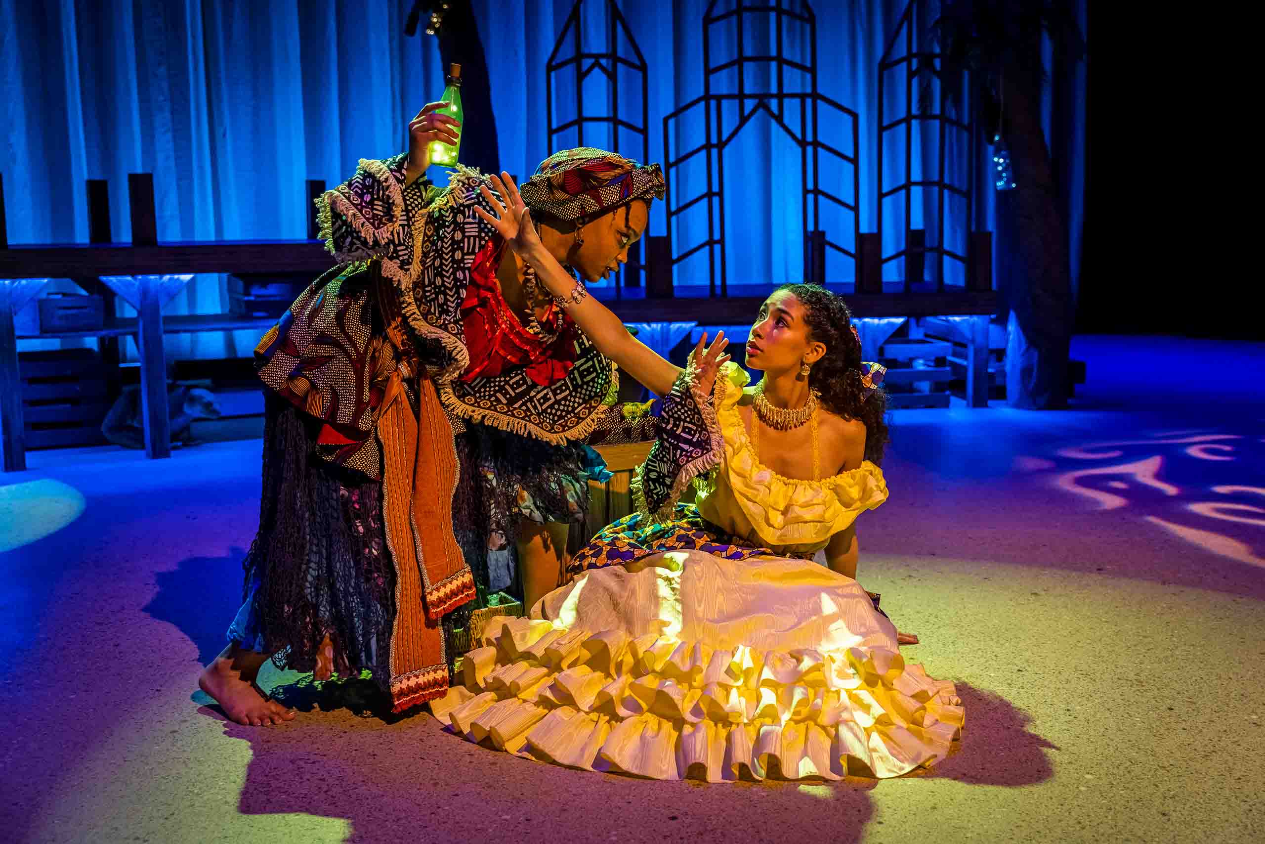 Two actors perform costumed in dresses with rich fabrics and ruffles, focused on a green bottle in the hand of one. An abstract set shines with blue and patterned lighting.
