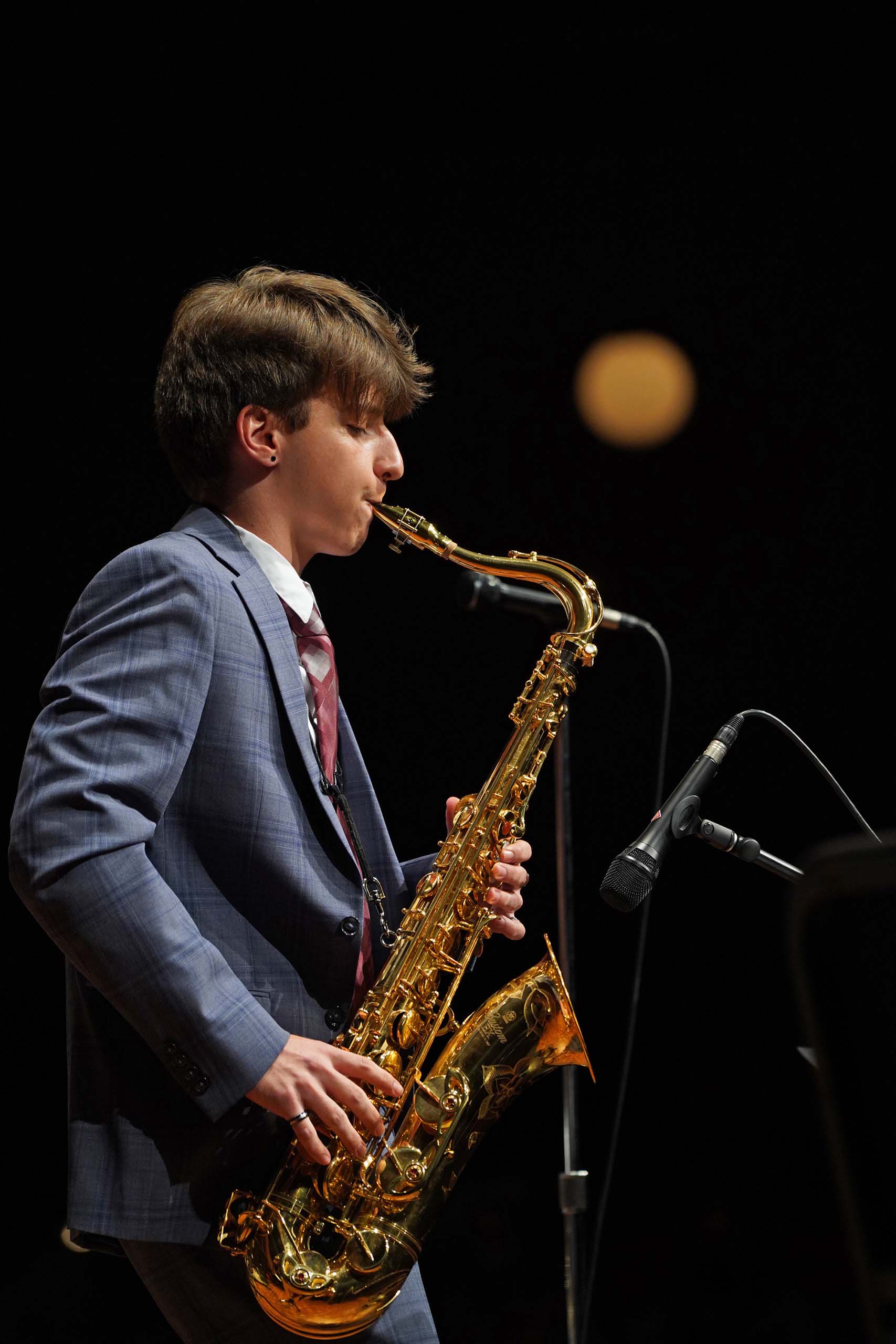 Anton Greene performs standing on saxophone, attired in a suit.
