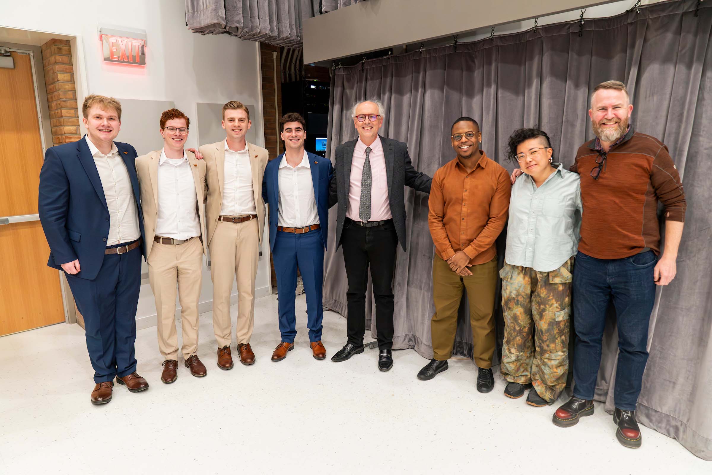 Eight people pose standing together in a rehearsal hall space.