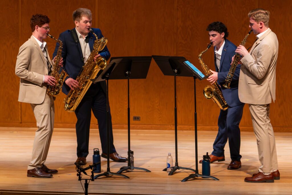 A saxophone quartet performs standing on stage with music stands and water bottles. Two are attired in blue suits, two are attired in light brown suits.