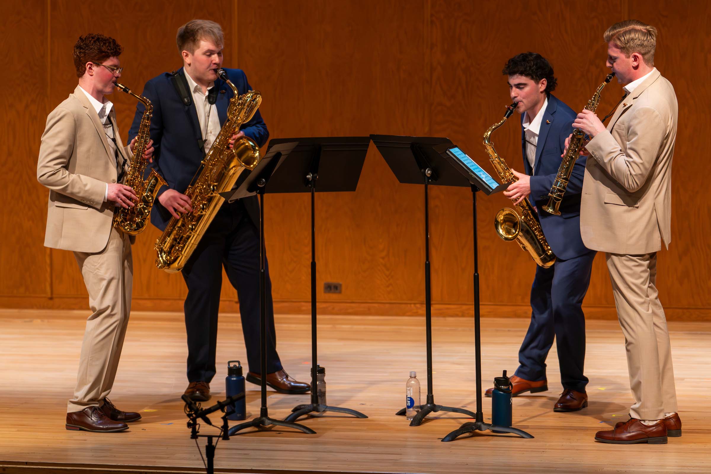A saxophone quartet performs standing on stage with music stands and water bottles. Two are attired in blue suits, two are attired in light brown suits.
