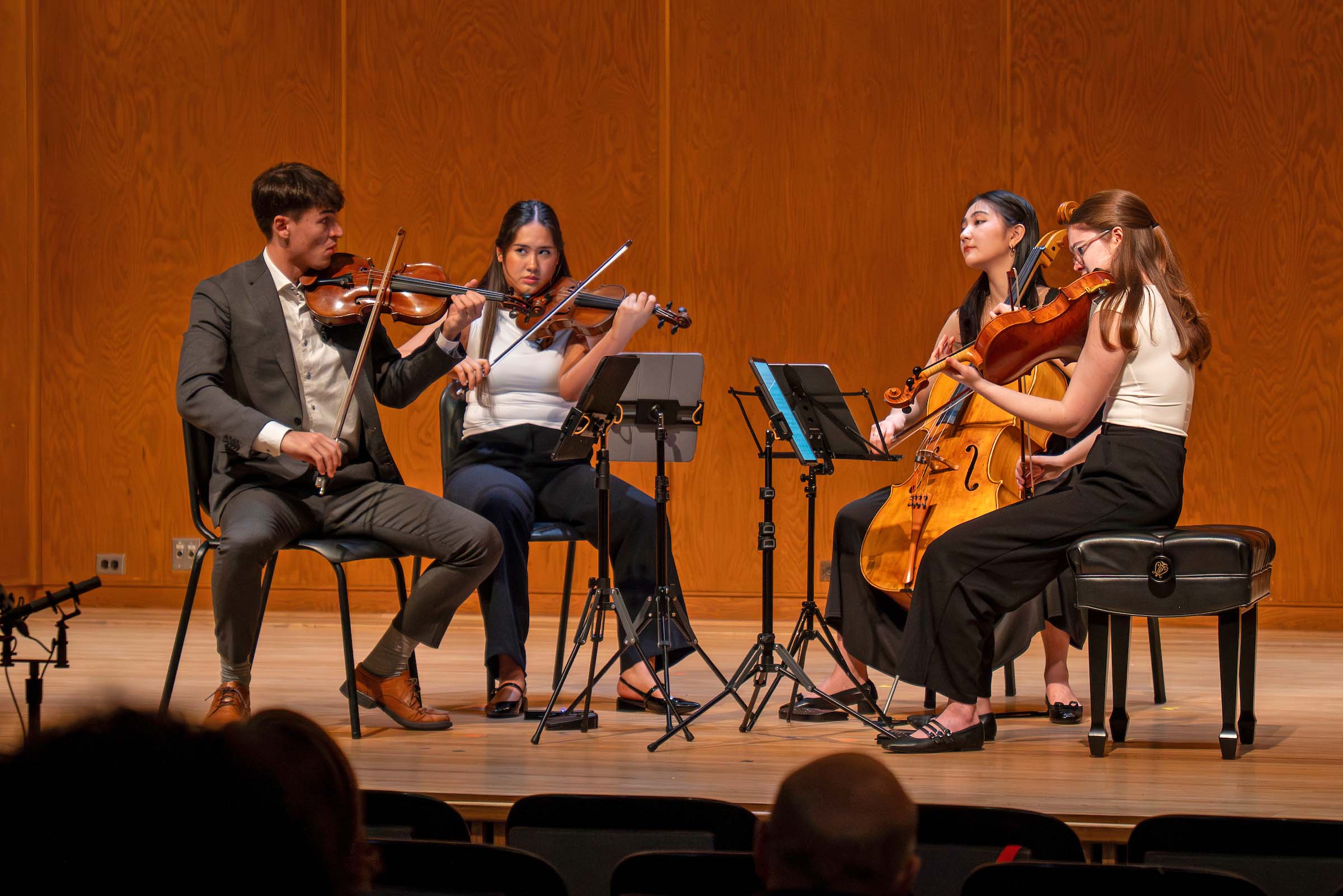 A string quartet performs seated on stage, with some audience heads visible in the foreground. All are wearing black and white concert attire.