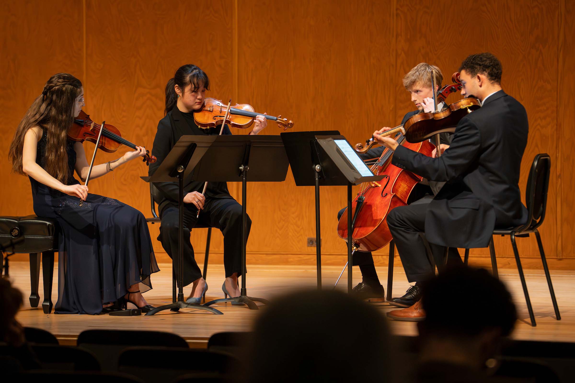 A string quartet performs seated on stage with music stands; some audience heads are visible in the foreground. All are wearing dark navy or black formal attire.