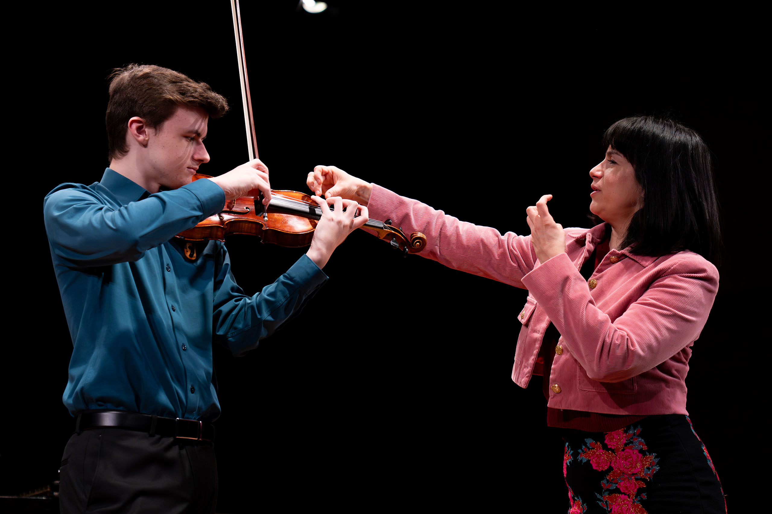 Standing on a stage in a performance, Danielle Belen works with a student playing violin.