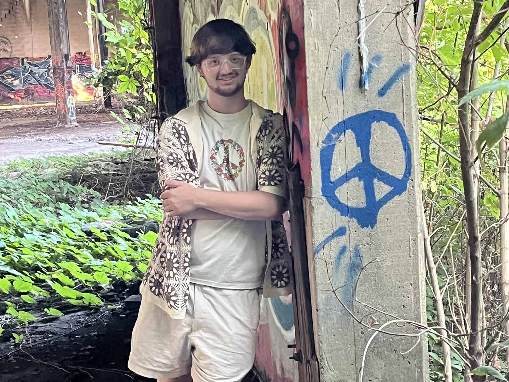 Christian Bush poses standing outdoors near concrete structures with graffiti, surrounded by foliage.