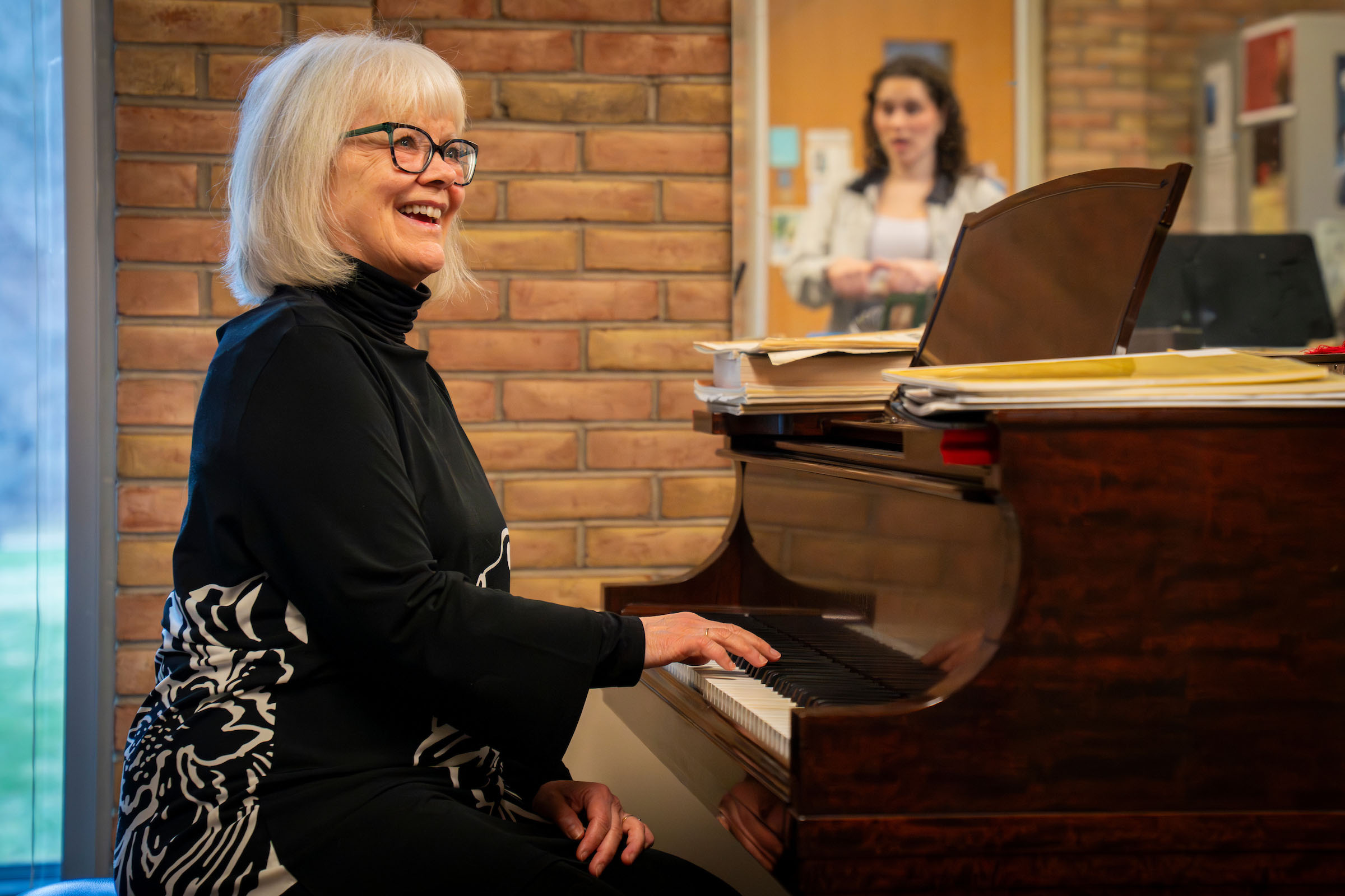 Freda Herseth plays a piano, watching a student sing whose reflection is seen in a mirror.