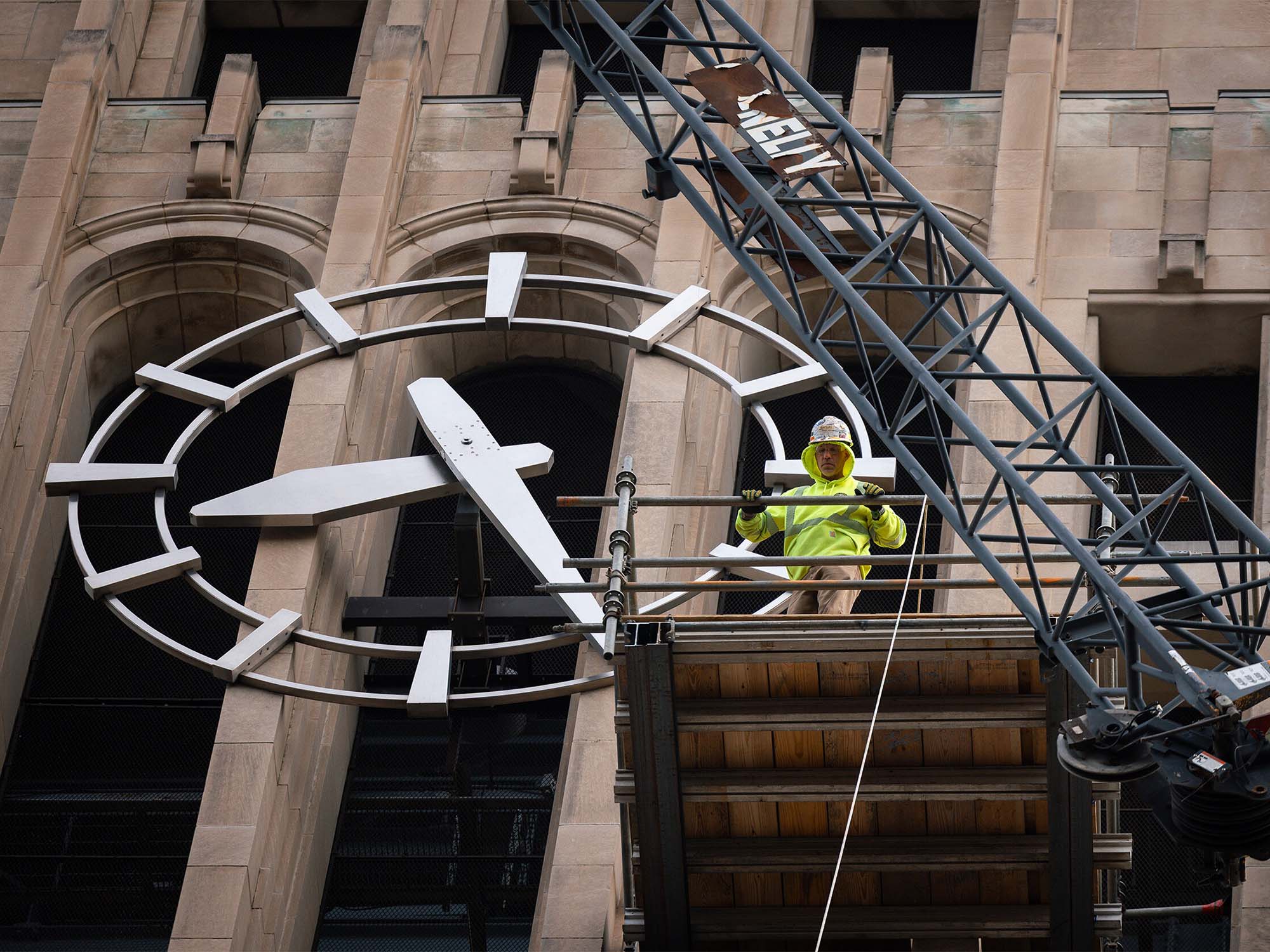 Near the clock face on the exterior of Burton Tower, a construction worker wearing bright green looks down from a hanging platform attached to a crane.