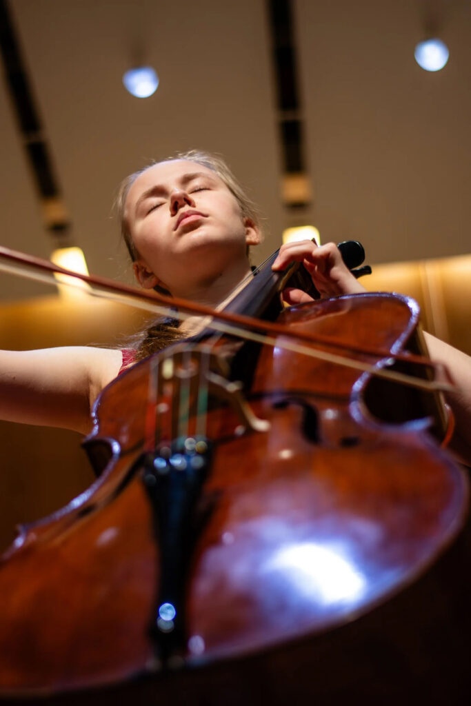 A young cellist performs in a hall, with a dramatic angled view from below the cello looking up.