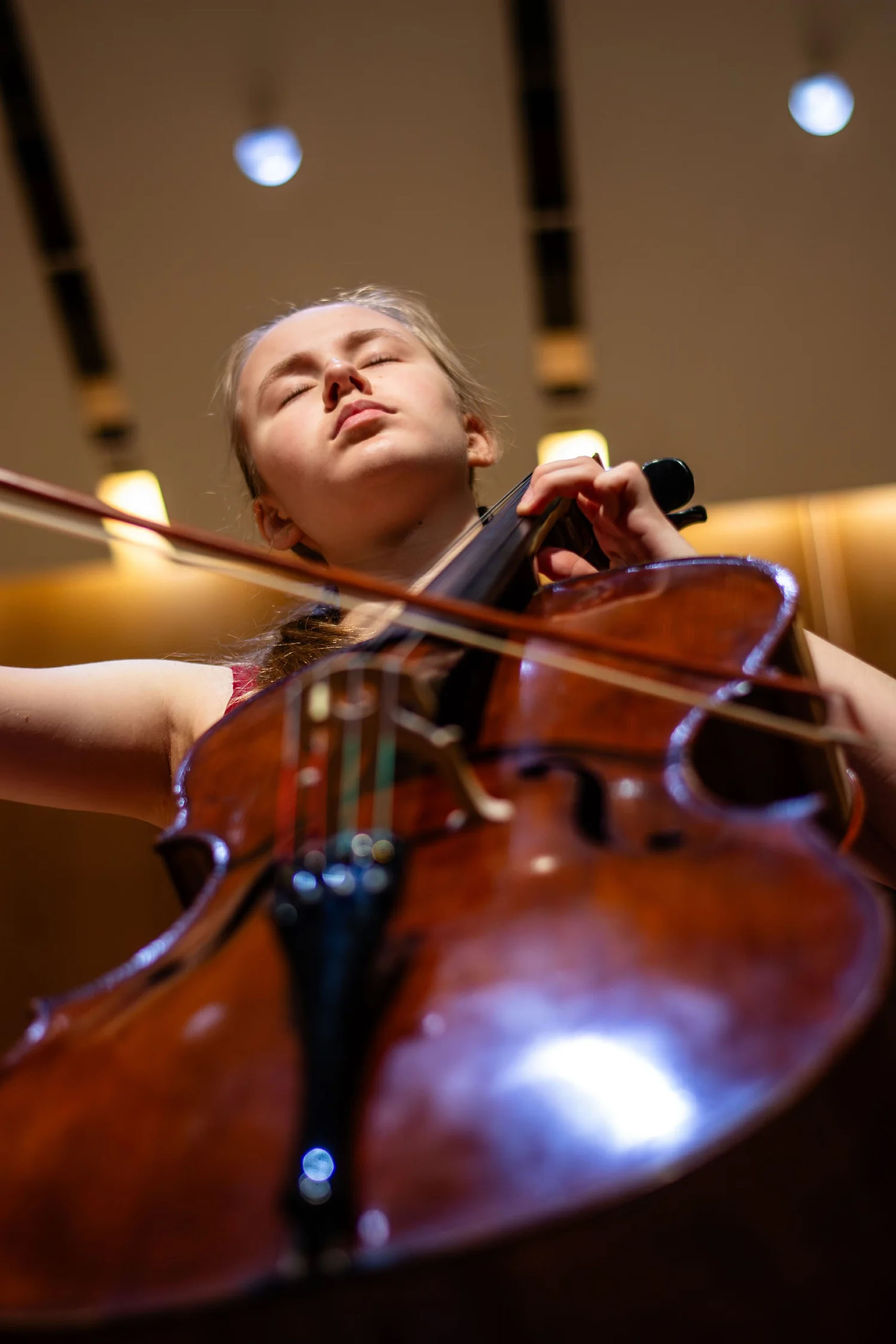 A young cellist performs in a hall, with a dramatic angled view from below the cello looking up.