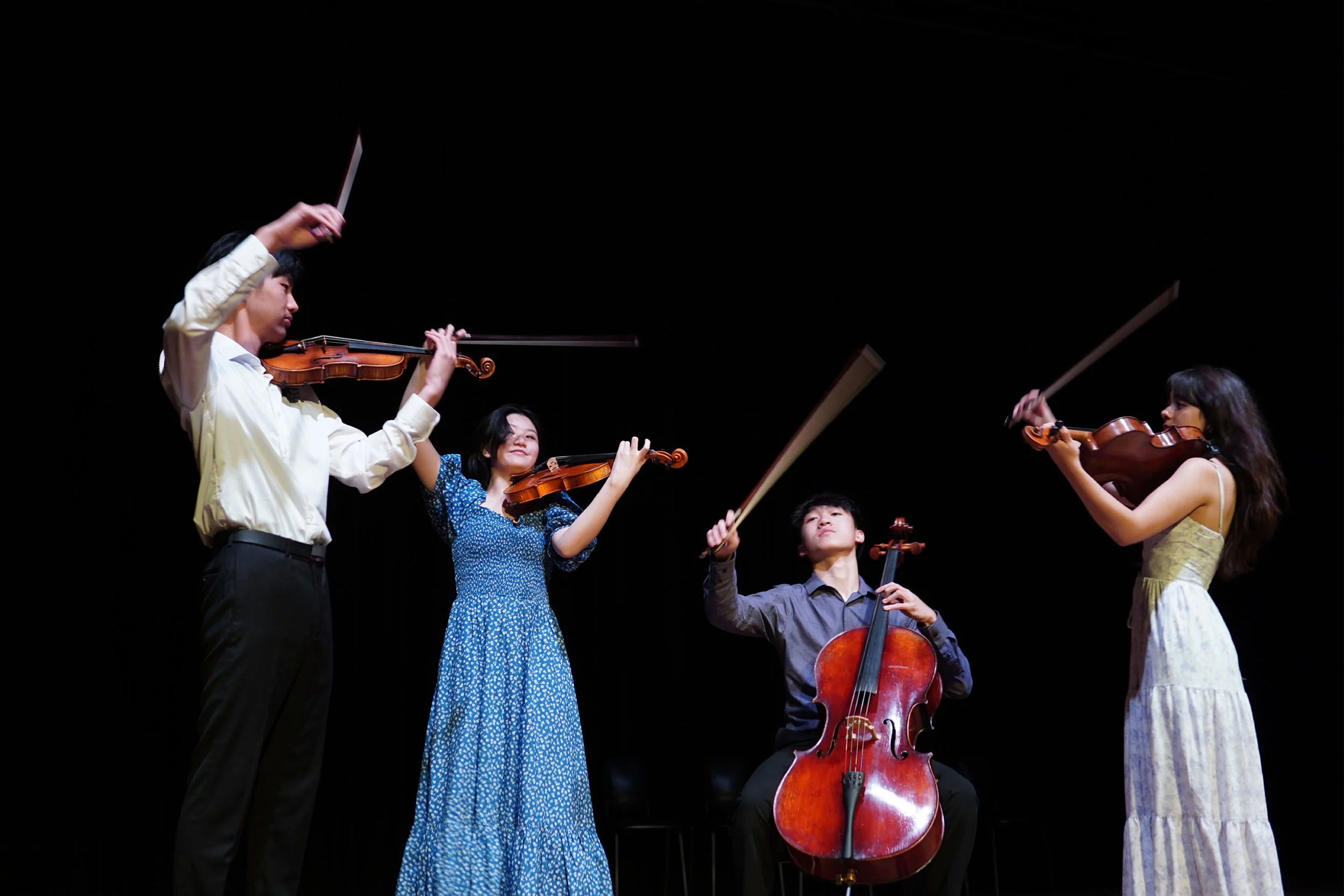 A string quartet of young performers play together on a dark stage, with their bows raised.