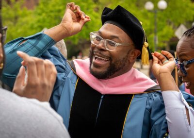 A doctoral graduate in academic regalia smiles in conversation with guests while standing outside.