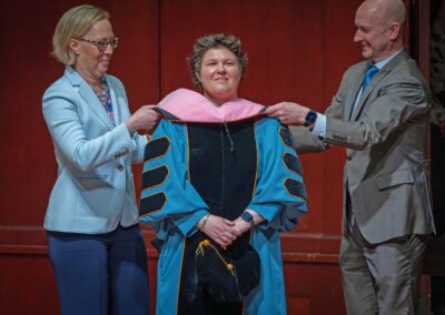 Faculty members Courtney Snyder and Jason Fettig place a pink hood over the shoulders of a doctoral graduate attired in academic regalia.