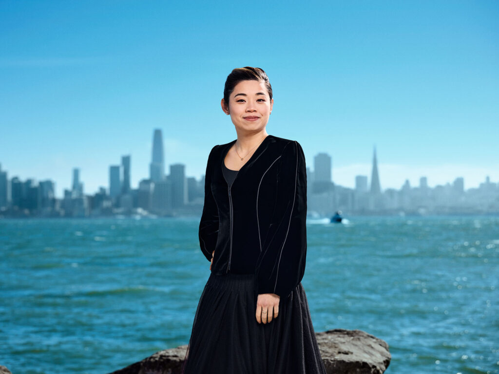 Portrait of Elim Chan standing outdoors next to the waters of the bay with the San Francisco skyline in the background.
